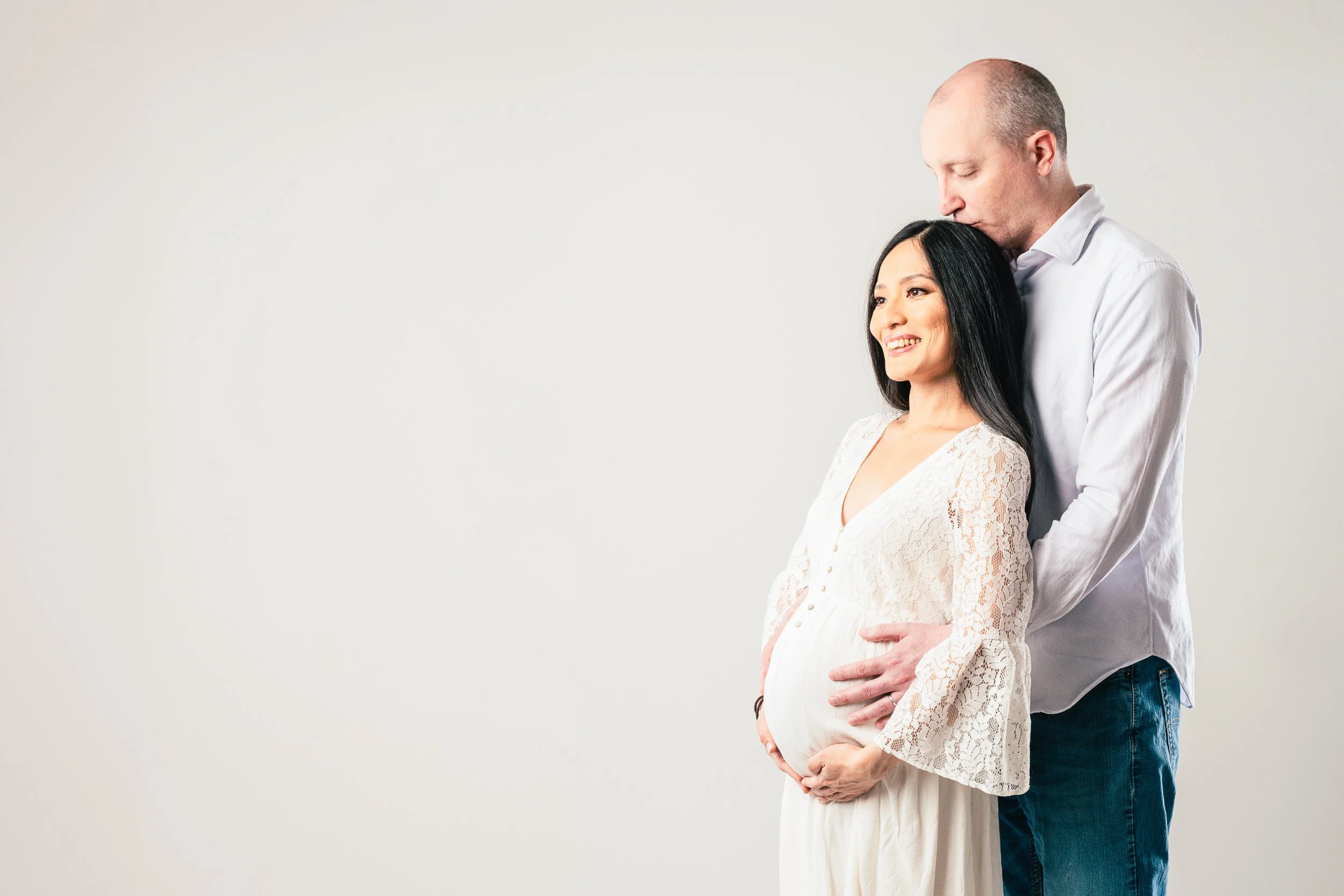 A pregnant woman with long black hair, wearing a white lace dress, smiling, with her partner standing behind her, gently holding her belly, and kissing her on the head.