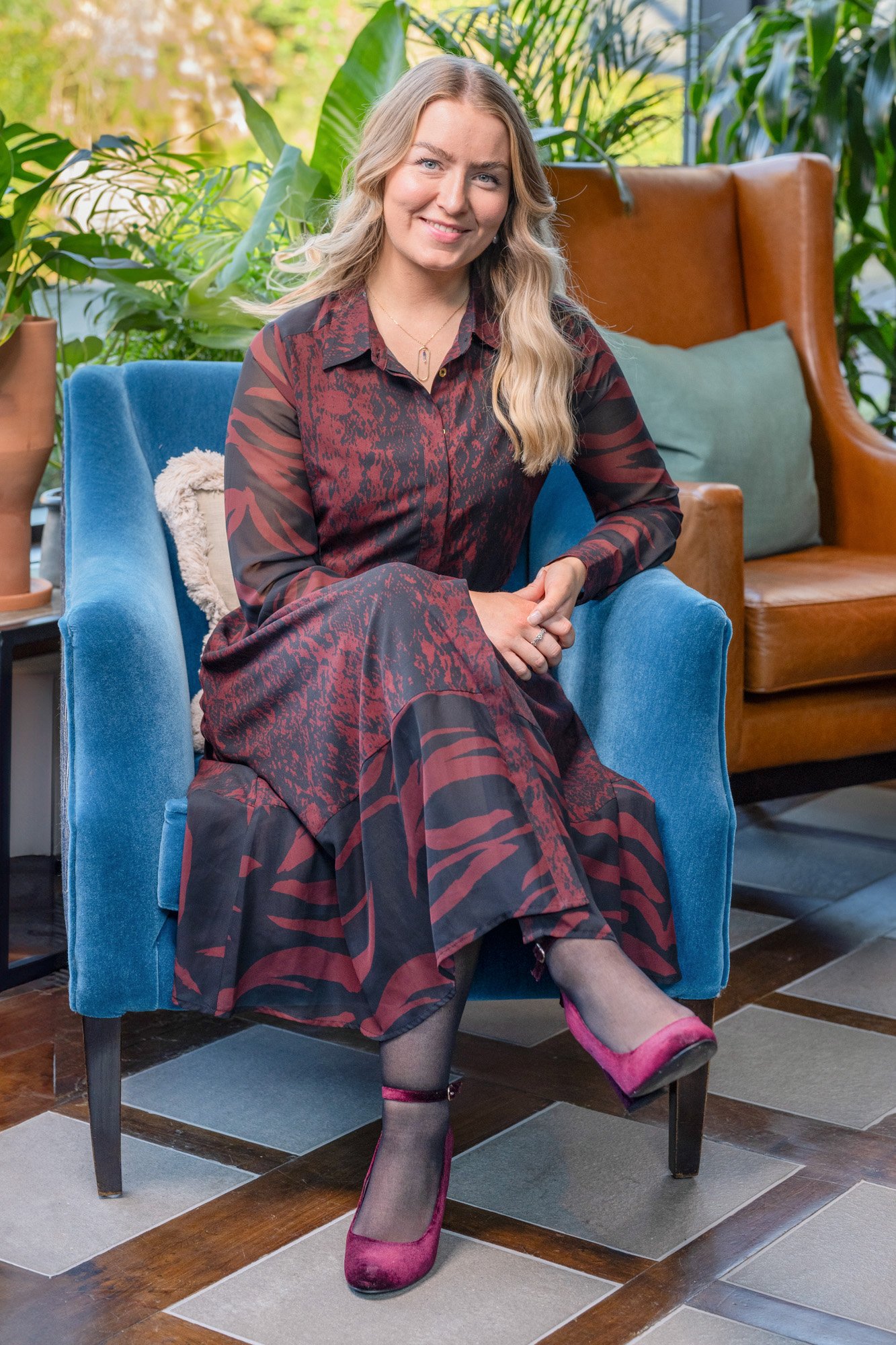 A woman with blonde wavy hair, smiling, sitting on a blue velvet armchair in a room with plants and natural light, wearing a long patterned dress and pink velvet heels.