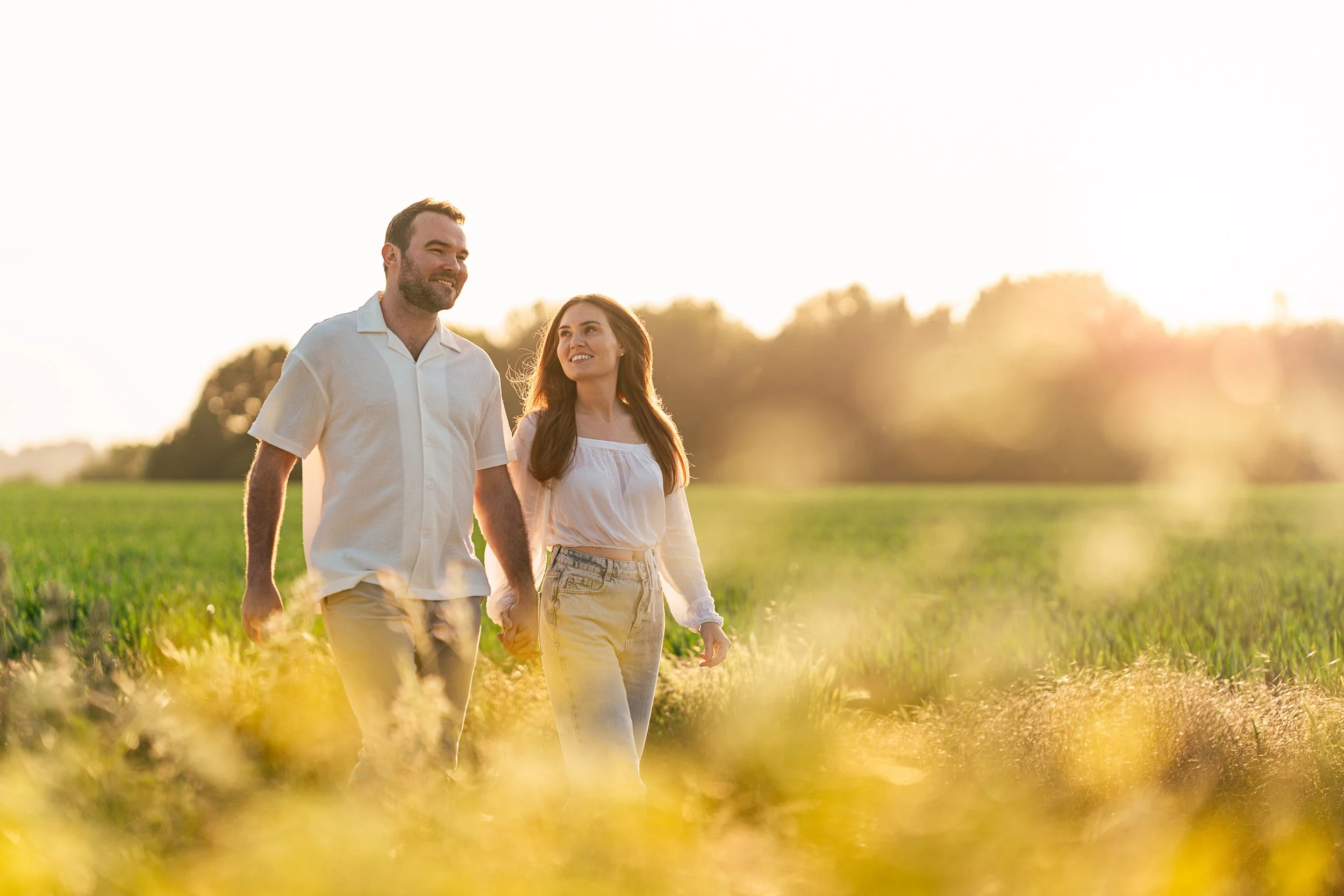 A happy couple walks hand-in-hand through a field during sunset.