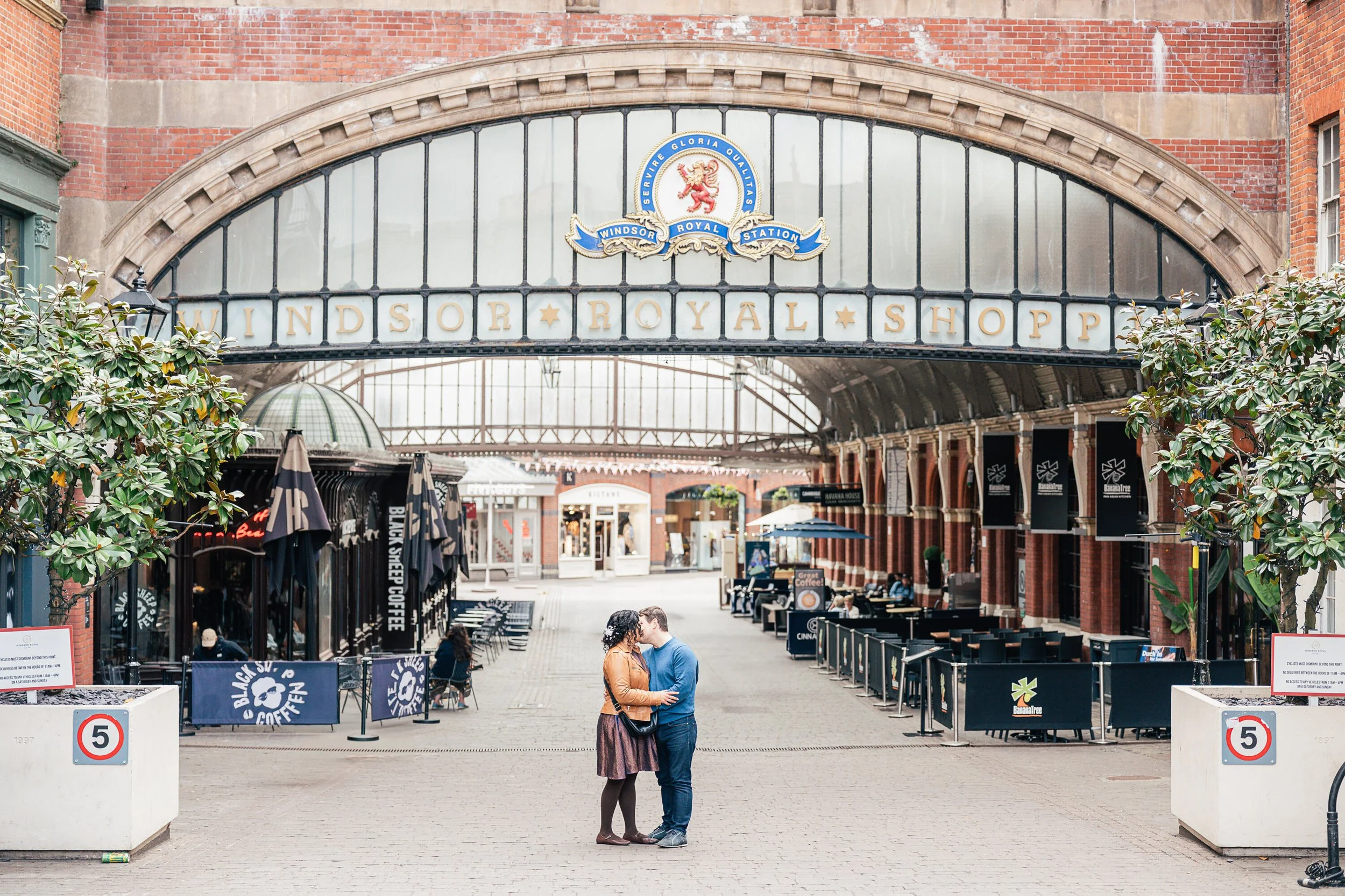 Two people standing close, kissing in front of Windsor Royal Station with a large archway on brick buildings, shops, outdoor seating, and trees around.