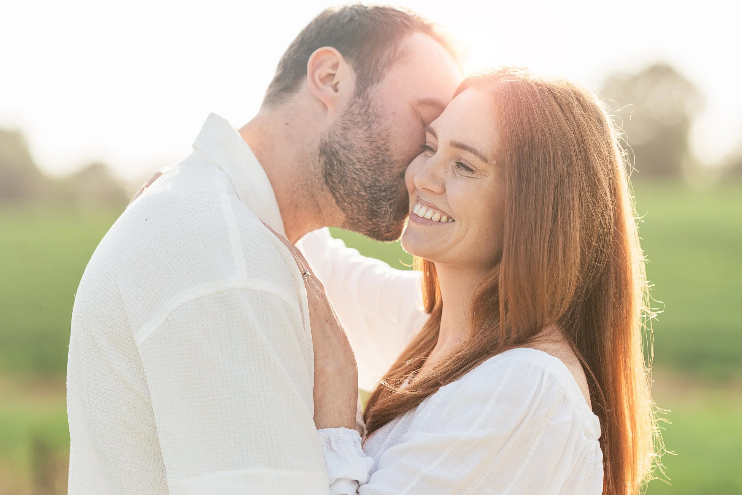 A couple embracing outdoors, with the man kissing the woman's forehead. The woman is smiling with her eyes closed, and they are standing in a field during sunset.