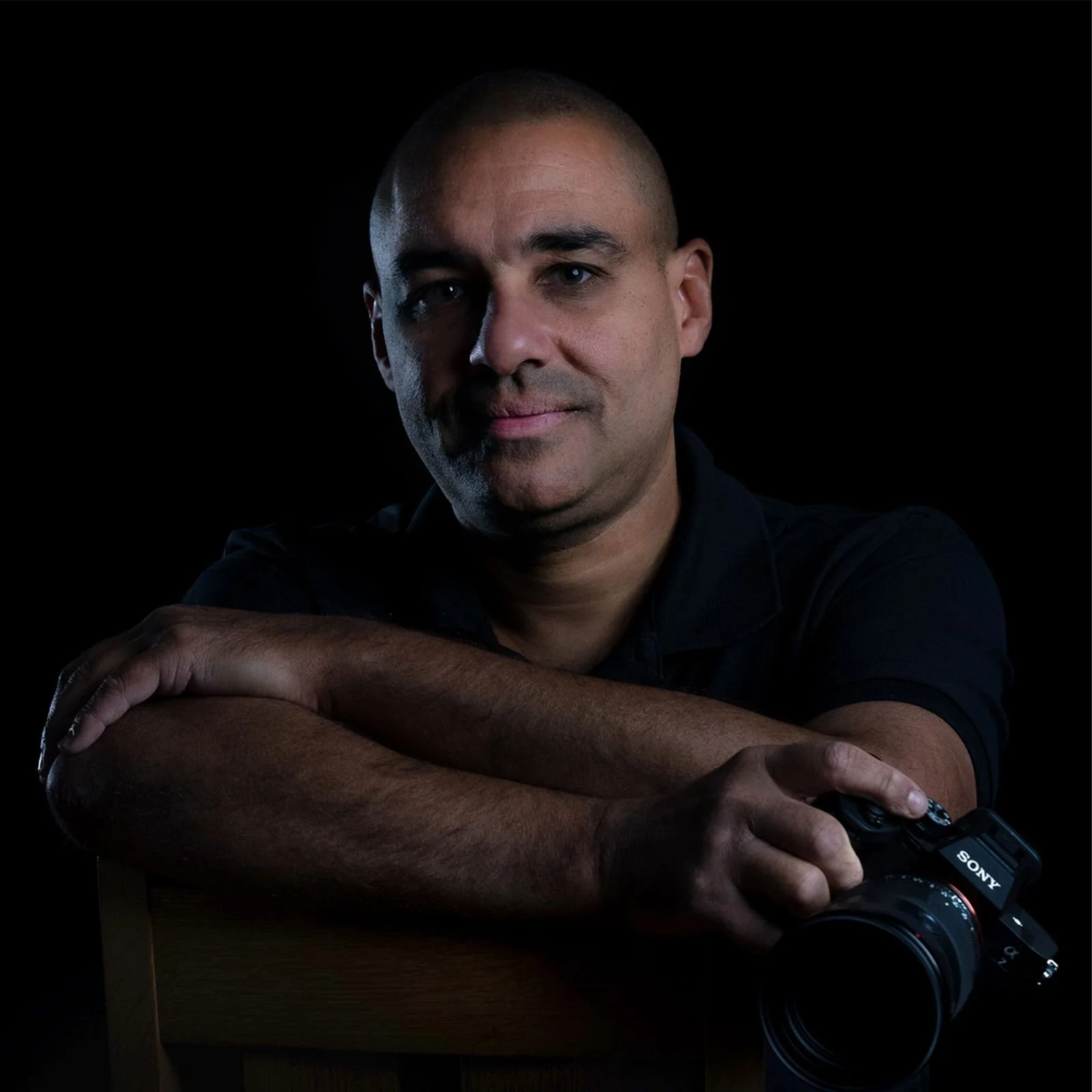 Portrait of a man with a shaved head holding a Sony camera, sitting with arms crossed on a wooden chair against a black background.