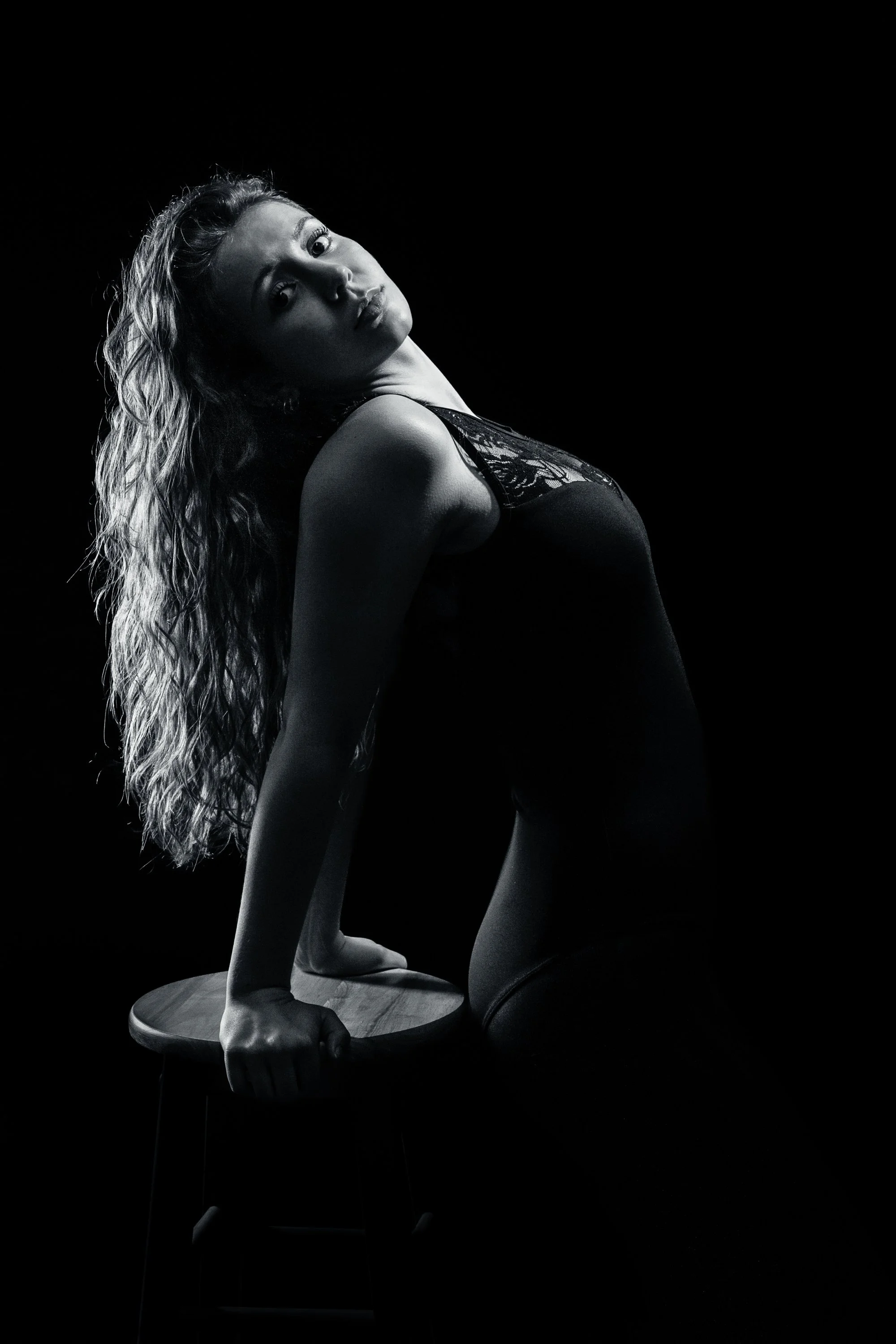 A black-and-white photo of a woman with long, curly hair posing on a stool, wearing a sleeveless dress, against a dark background.