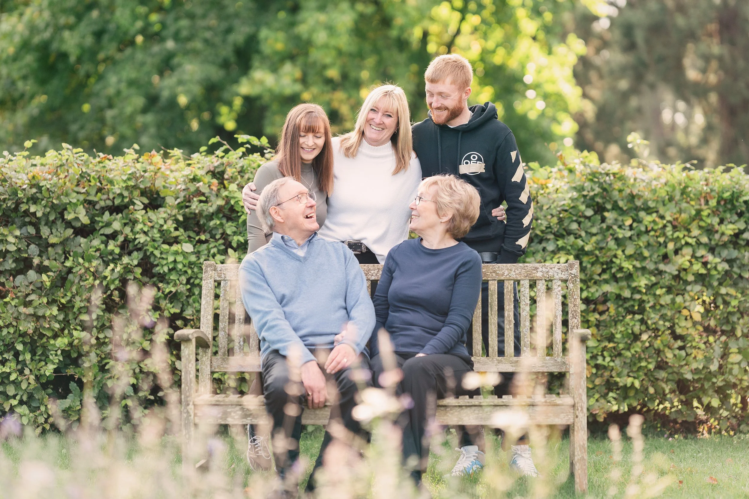 A multigenerational family of six people, including an elderly couple seated on a park bench and four younger adults standing behind them, smiling and enjoying a day outdoors with green trees and bushes in the background.