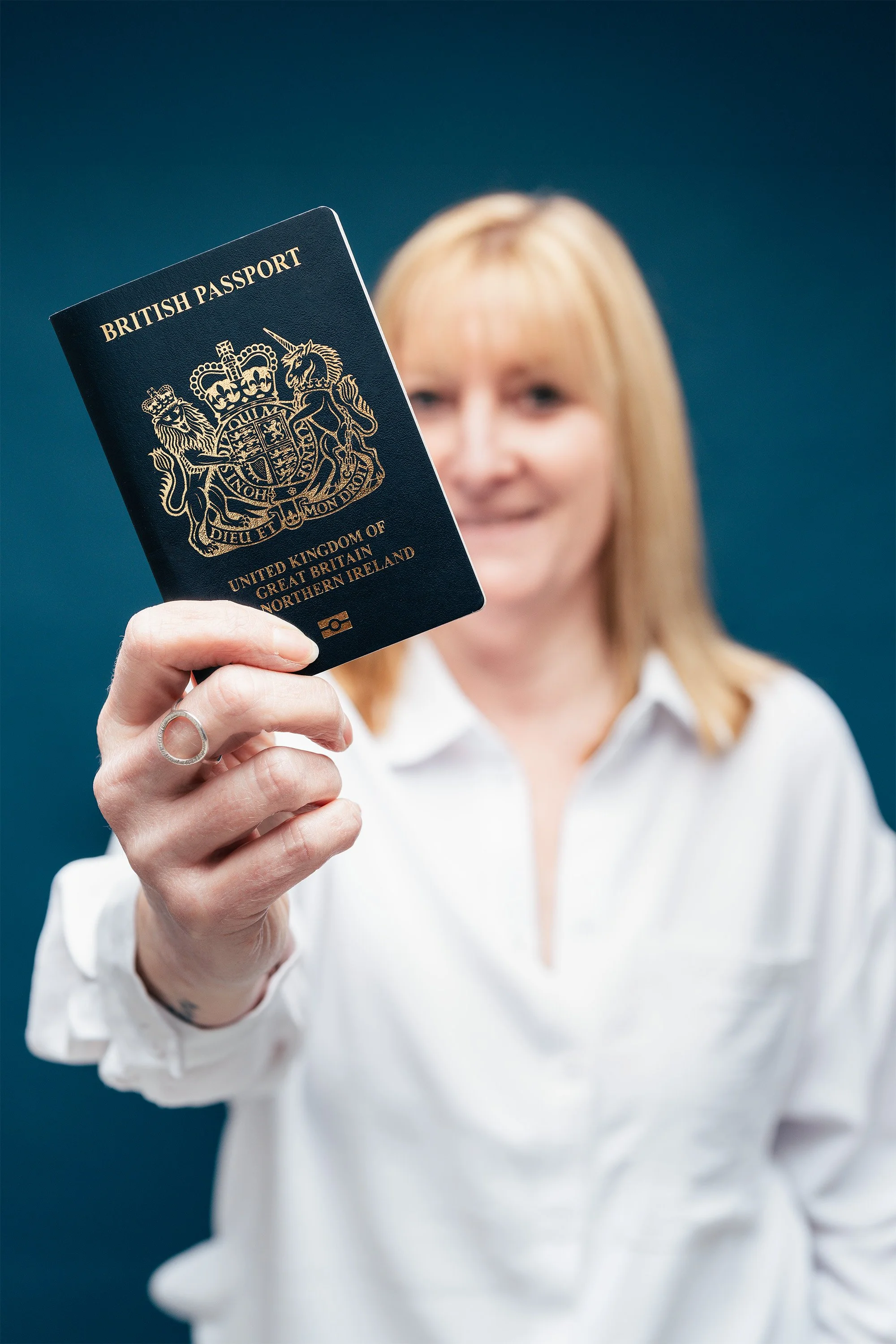 A woman with blonde hair and a white blouse holding a British passport up towards the camera, with a blue background.