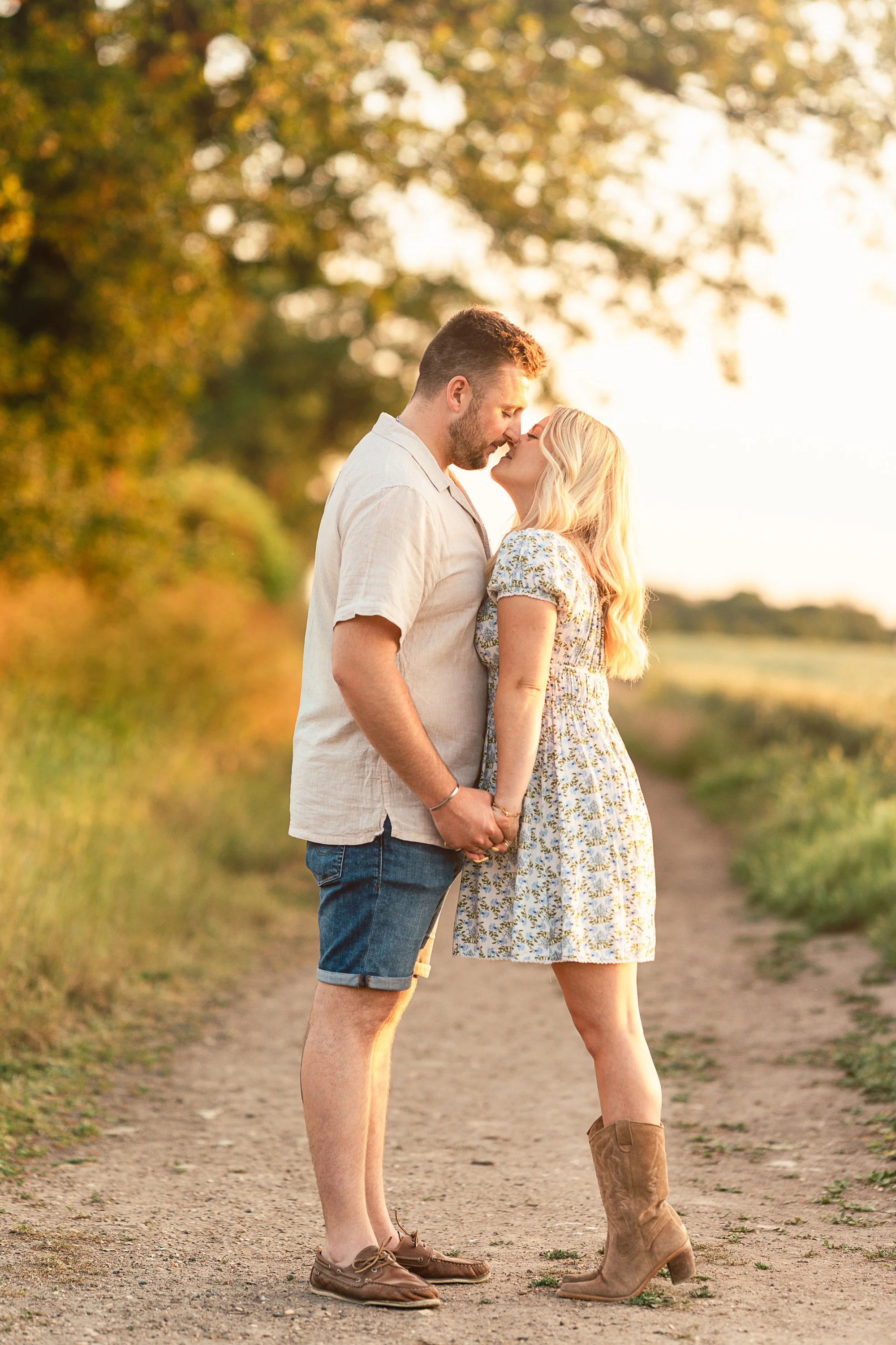 A couple stands closely on a dirt path, holding hands, with their foreheads touching and eyes closed, during golden hour in a rural outdoor setting.
