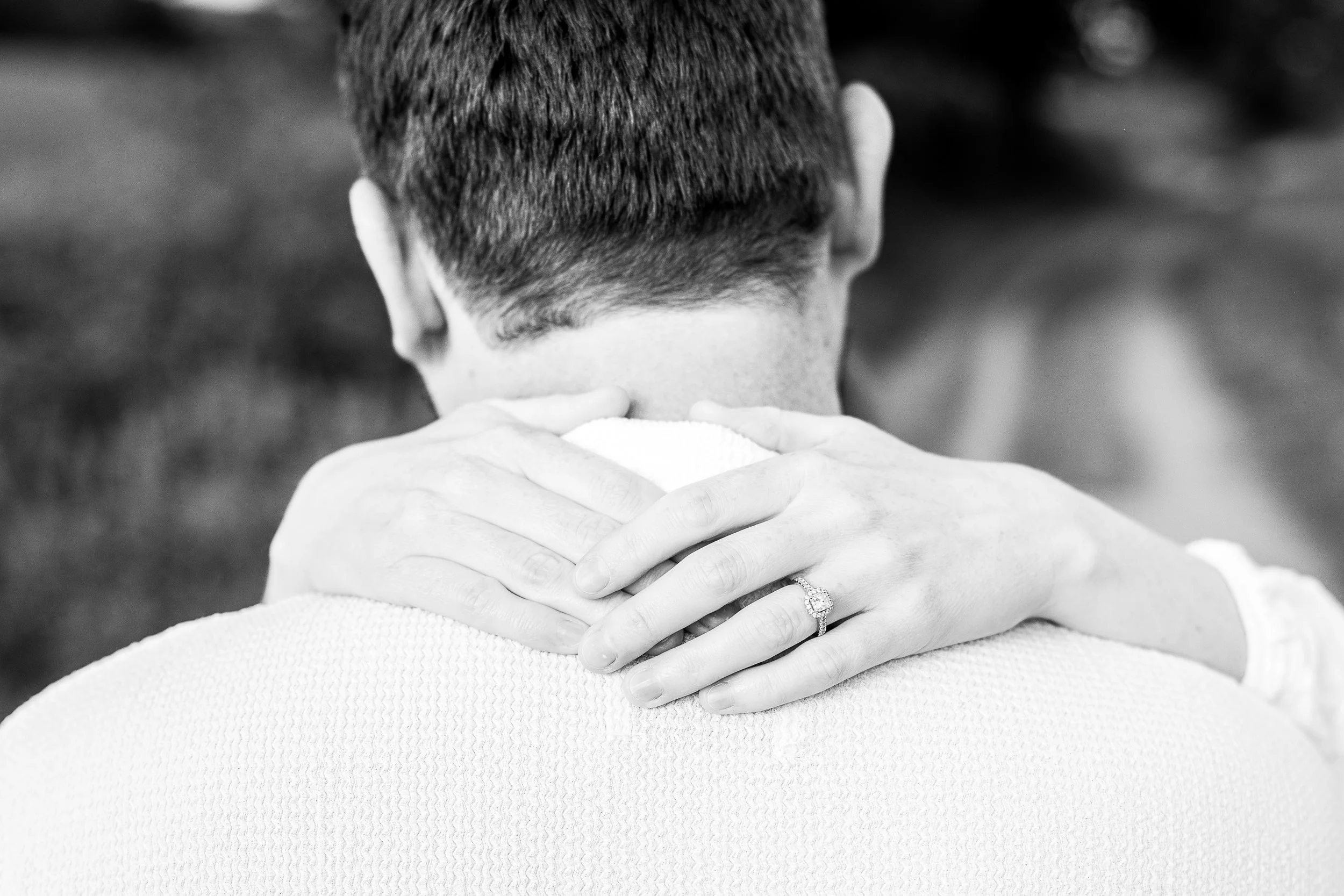 A black-and-white photo of a person with short hair, seen from behind, with their arms crossed over their shoulders. A woman’s hand with a ring is resting on the person’s shoulder.