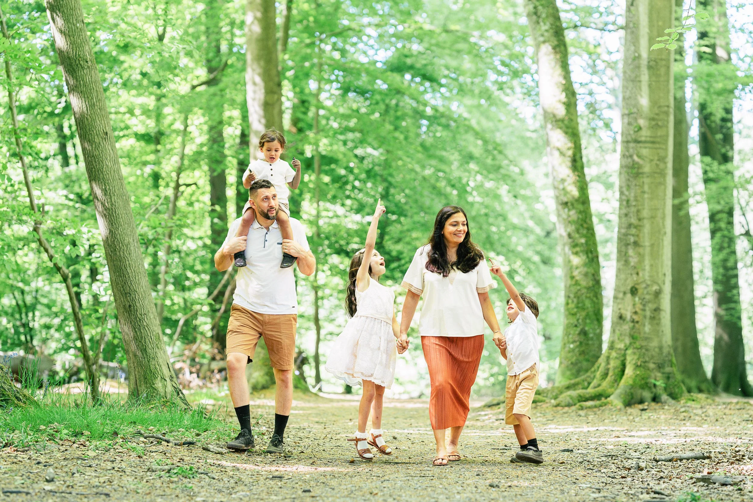 A family walking through a lush green forest. The man carries a young girl on his shoulders, while the woman holds hands with two children. The kids are pointing and reaching, enjoying their time outdoors.