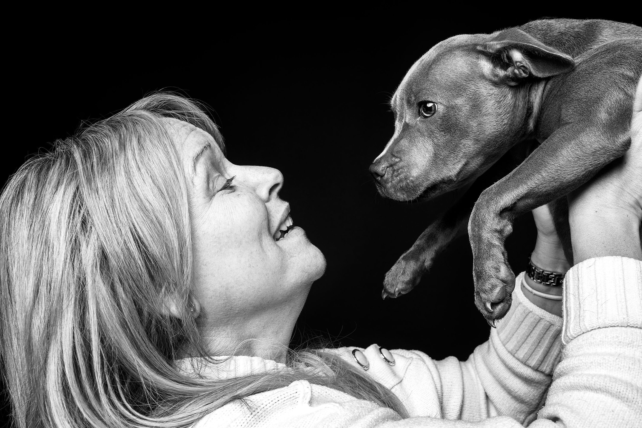 A woman smiles and looks at a puppy she is holding up in front of her, against a black background.