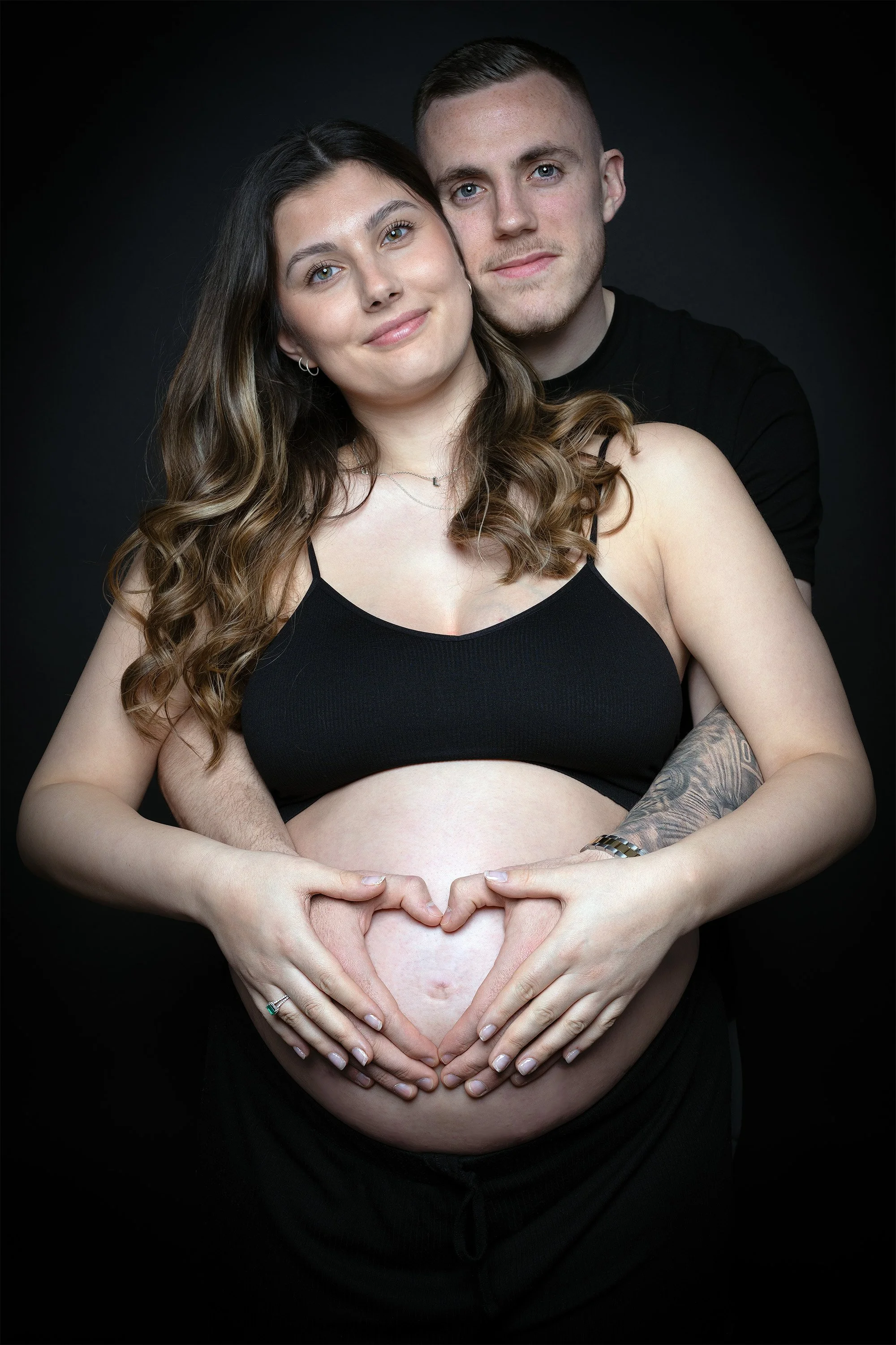 A pregnant woman and her partner posing together against a black background. The woman's belly is visible, and they form a heart shape with their hands over her abdomen.