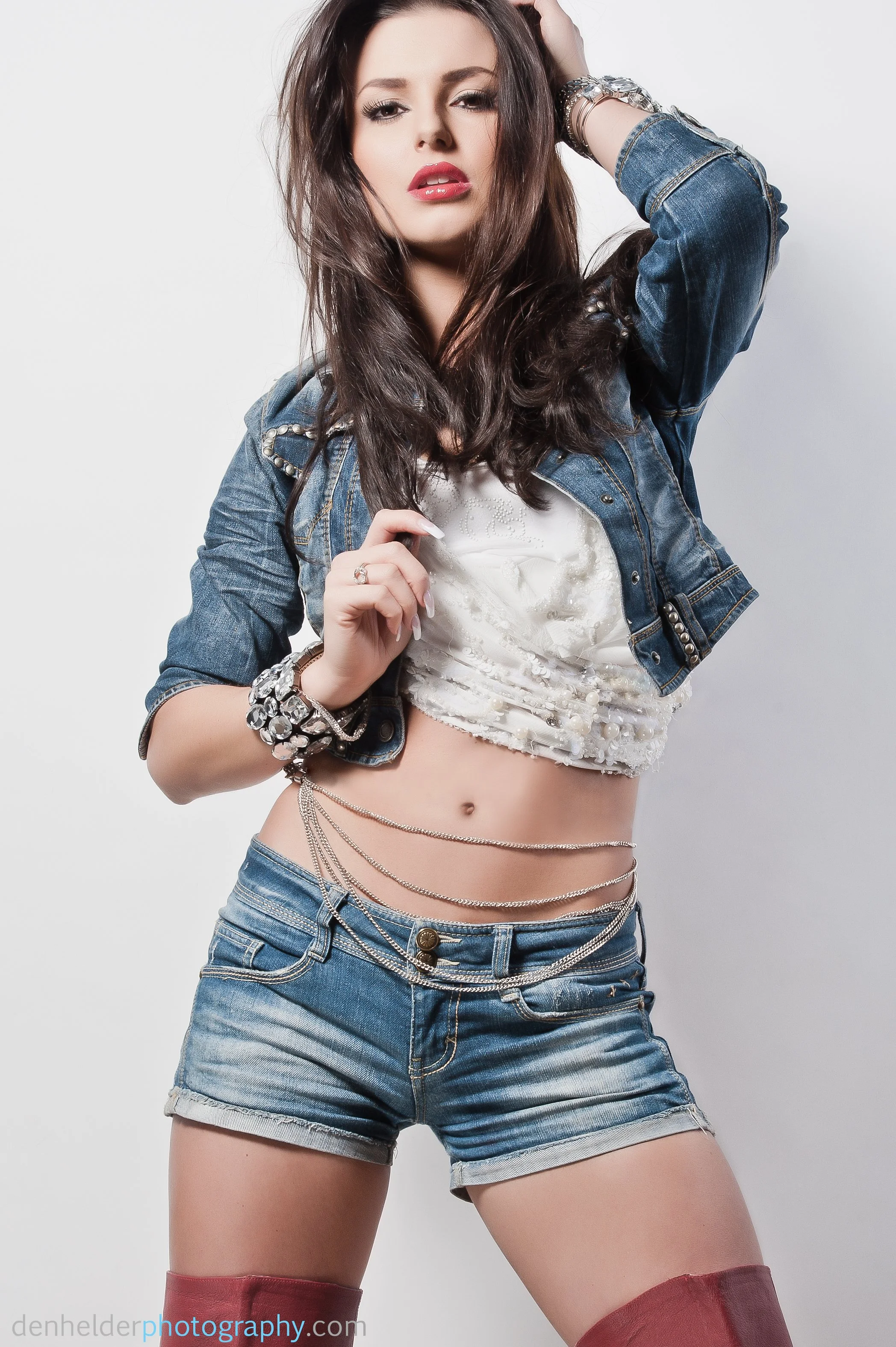 A young woman with long brown hair, wearing a denim jacket, white crop top, denim shorts, red thigh-high stockings, and jewelry, posing against a plain white background.