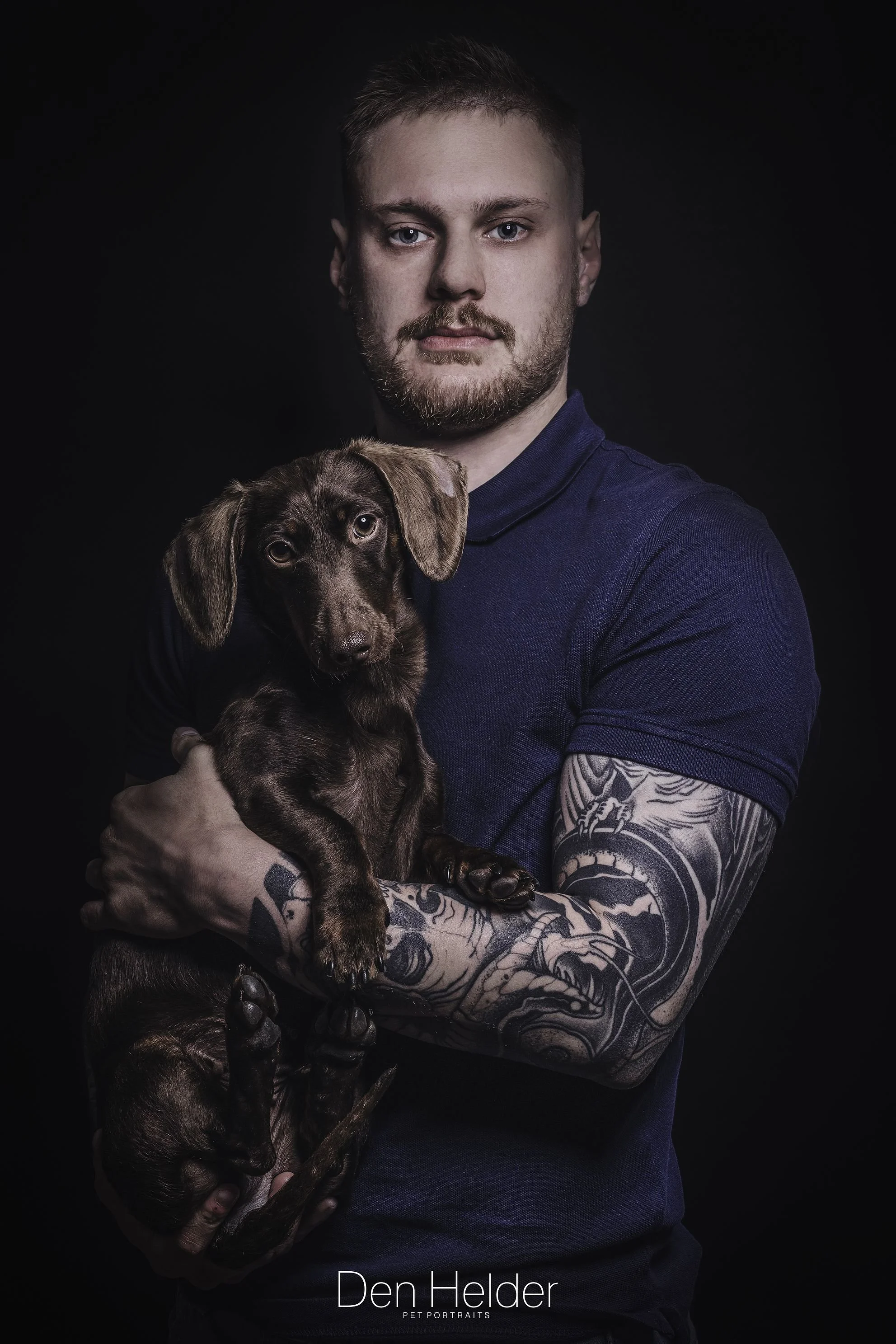 A man with tattoos on his arms holding a brown dachshund puppy against a dark background.