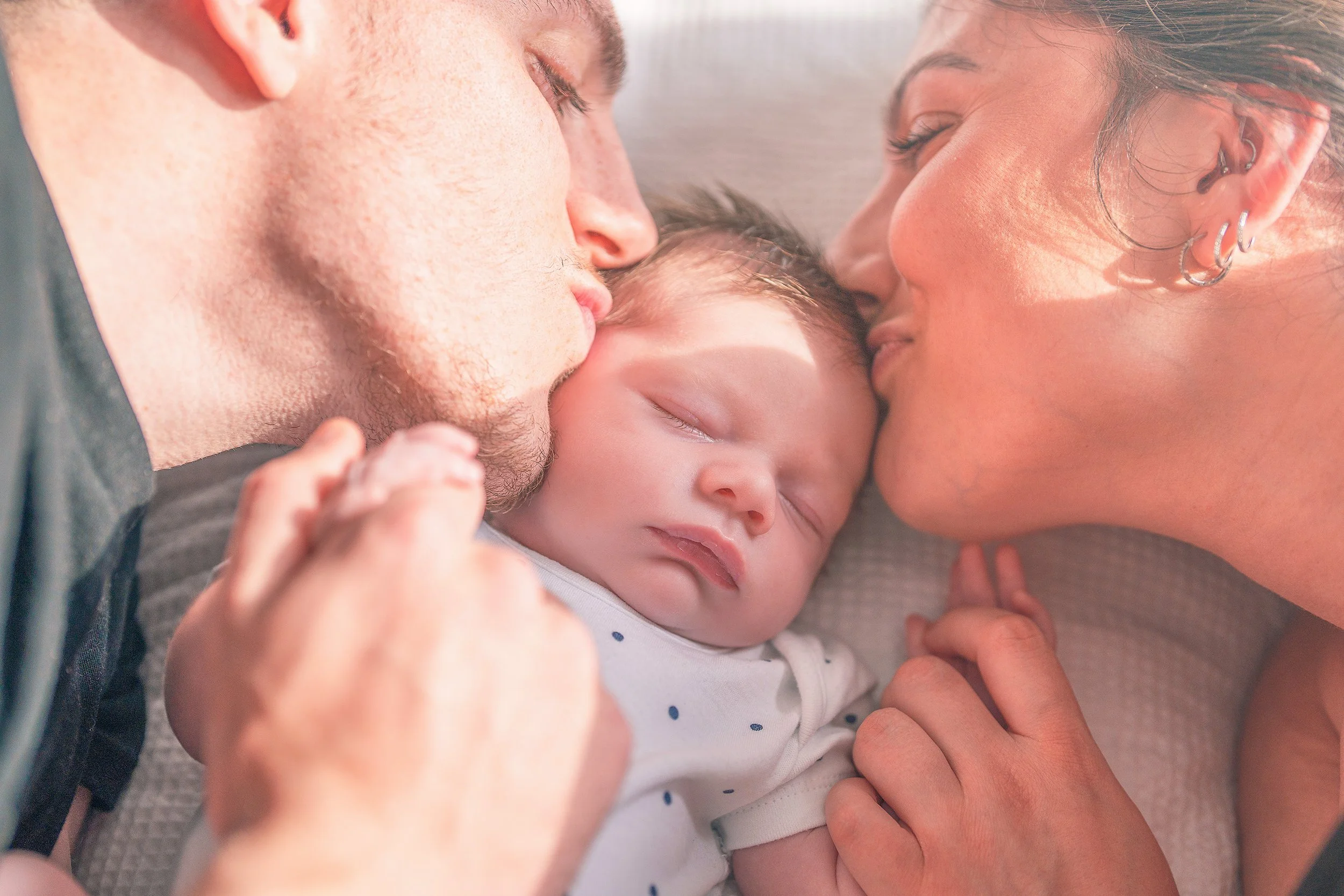 A baby sleeping peacefully between two adults, who are kissing the baby's cheeks.