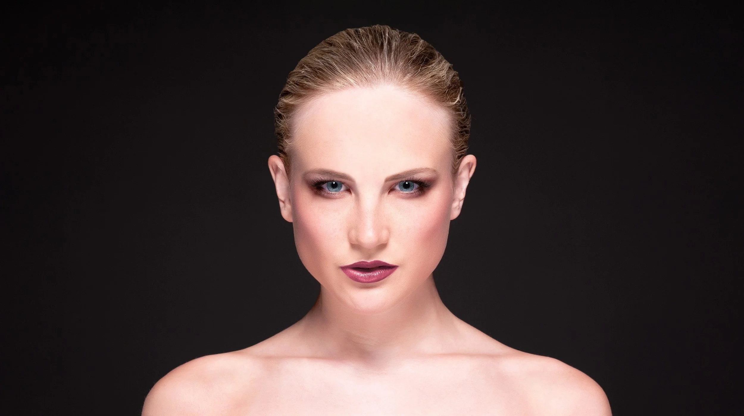 A close-up portrait of a woman with fair skin, blonde hair slicked back, blue eyes, wearing makeup, against a black background.