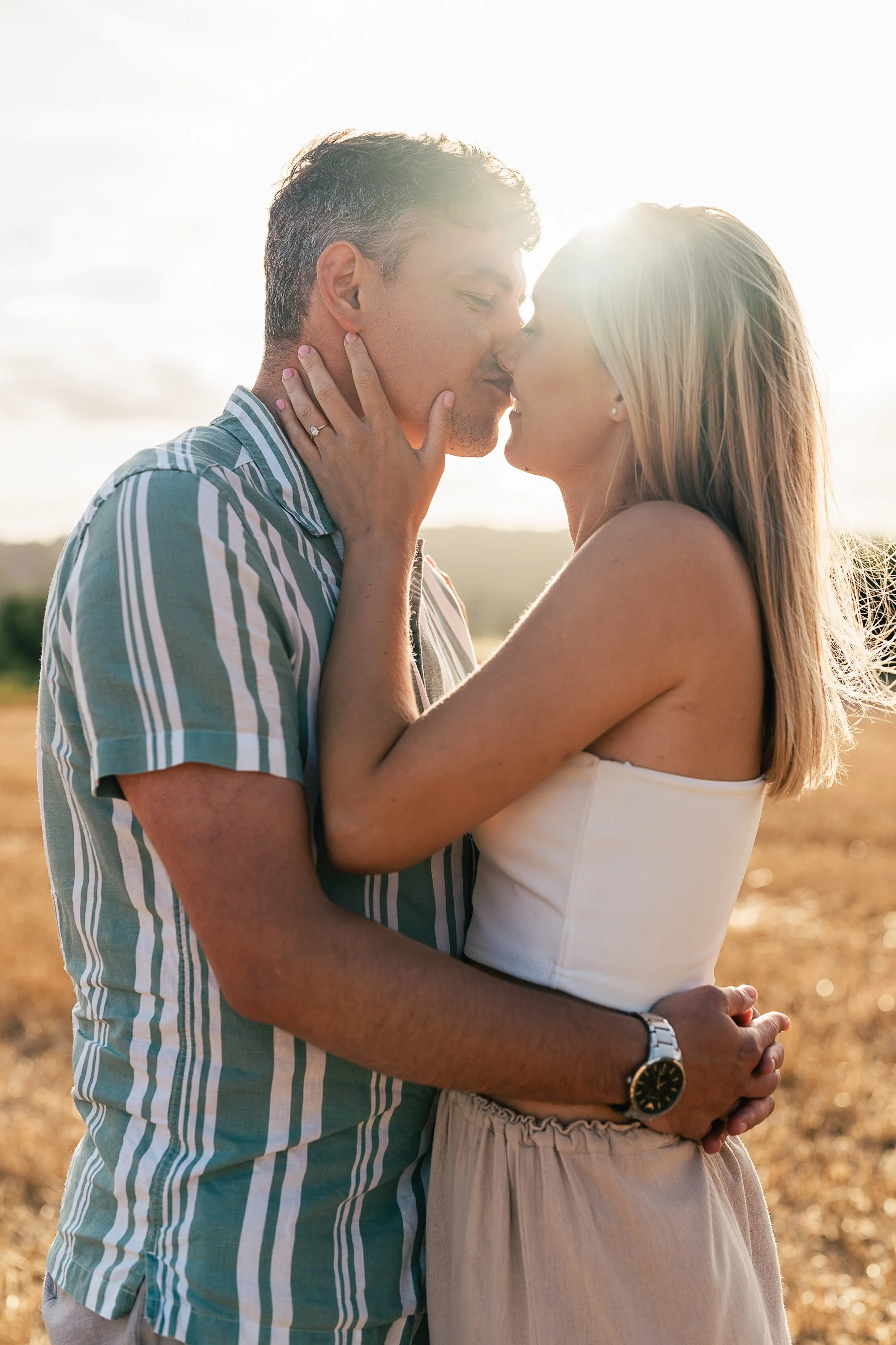 A couple kissing outdoors during sunset, with the woman holding the man's face and the man holding the woman's waist.