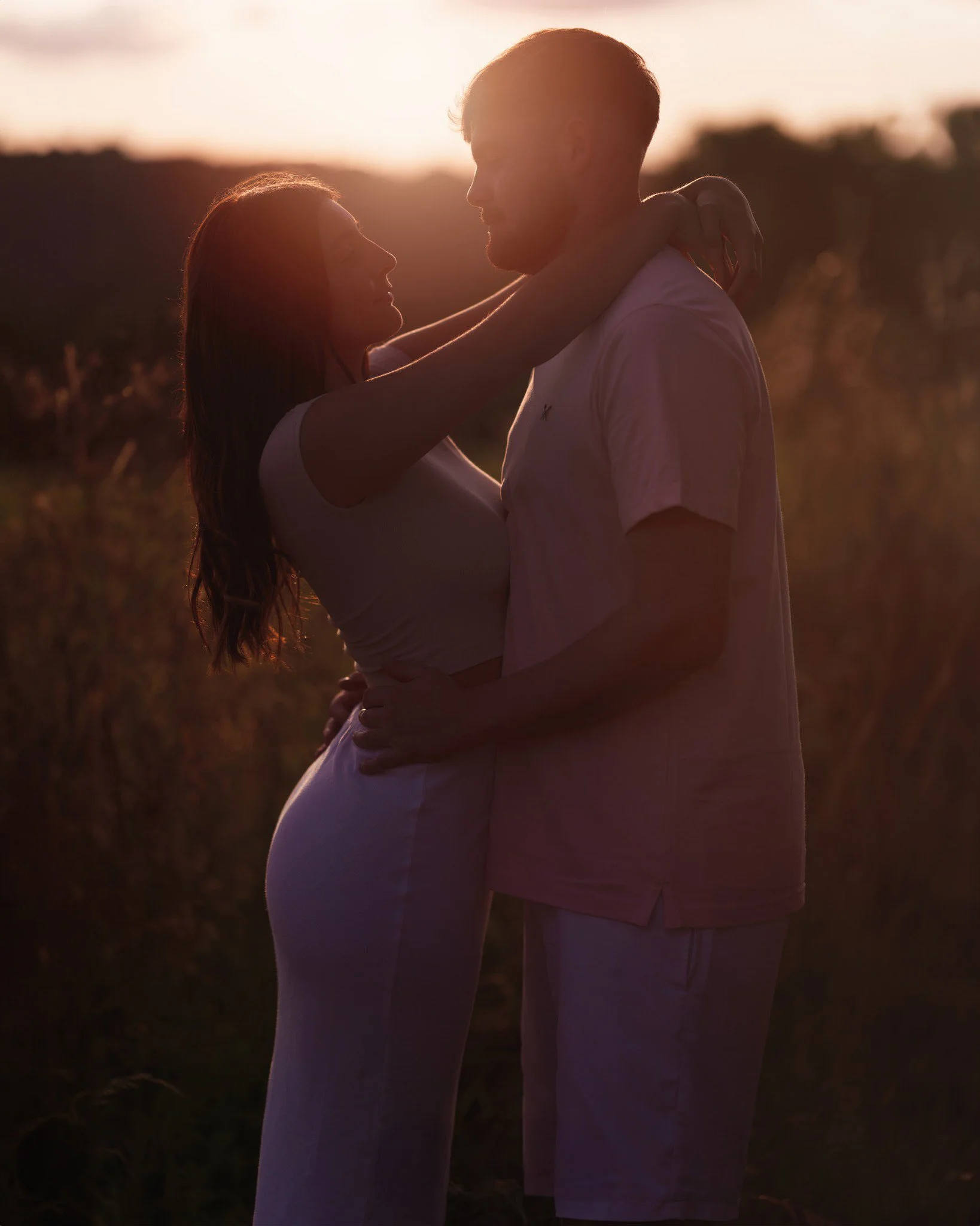A couple is embracing outdoors at sunset, with the woman’s arms around the man’s neck and the man’s hands on the woman’s waist, both gazing into each other's eyes.