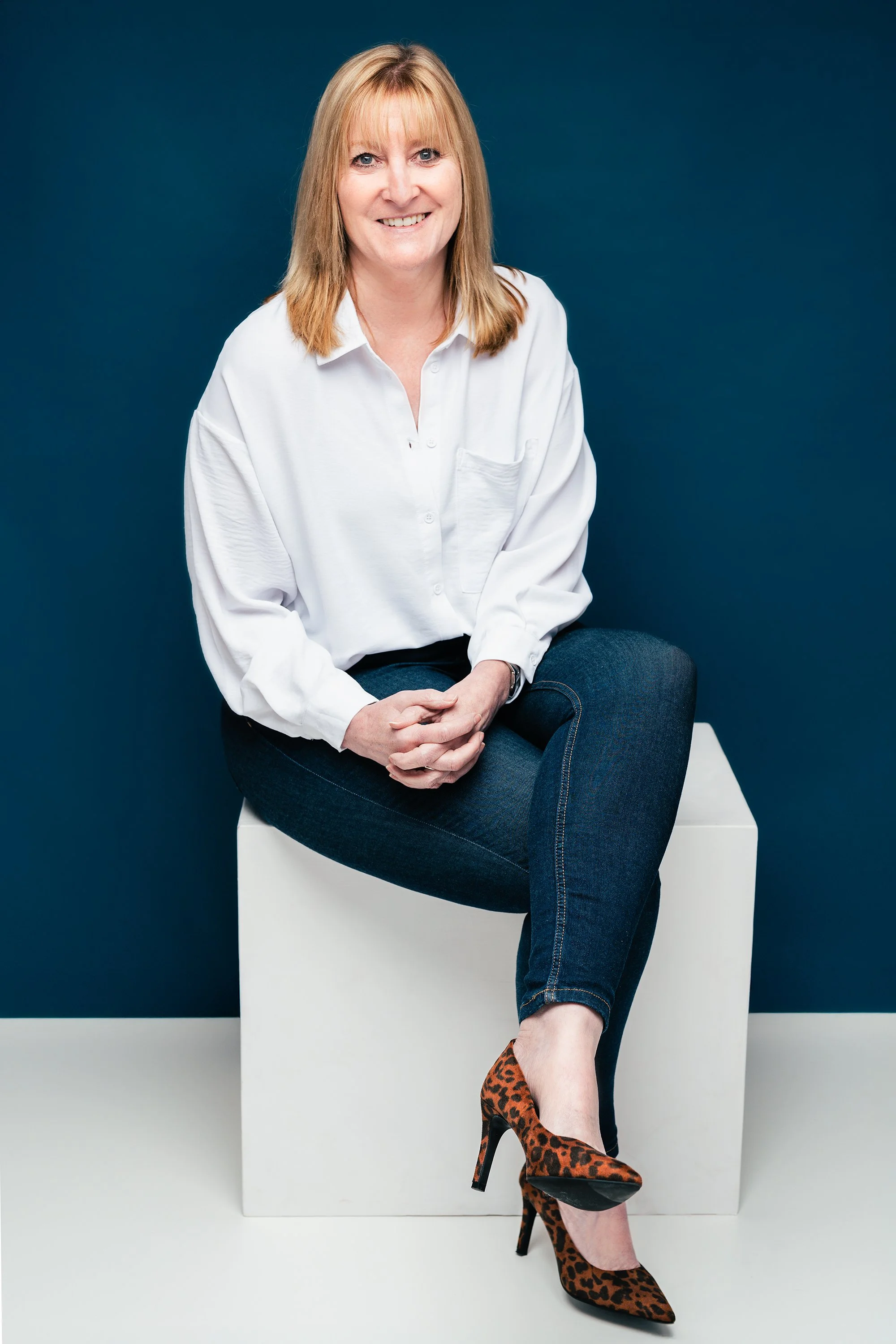 A woman with blonde hair wearing a white button-up shirt, dark blue jeans, and leopard print high heels, sitting on a white cube against a dark blue background.