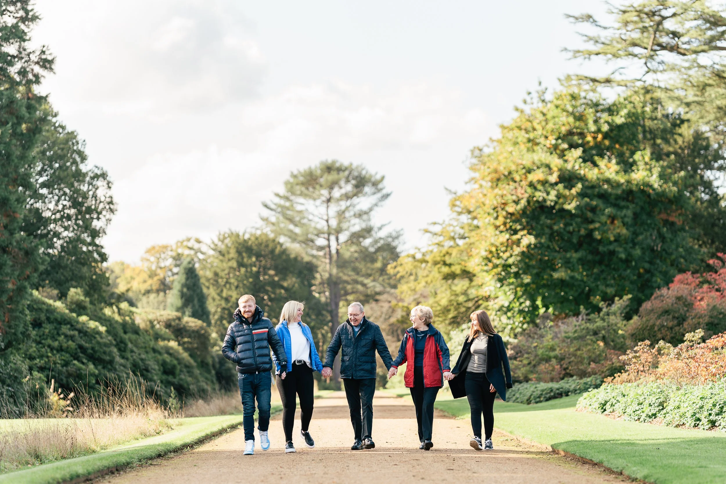 Five people walking hand in hand along a park pathway during fall or spring, surrounded by colorful trees and greenery.