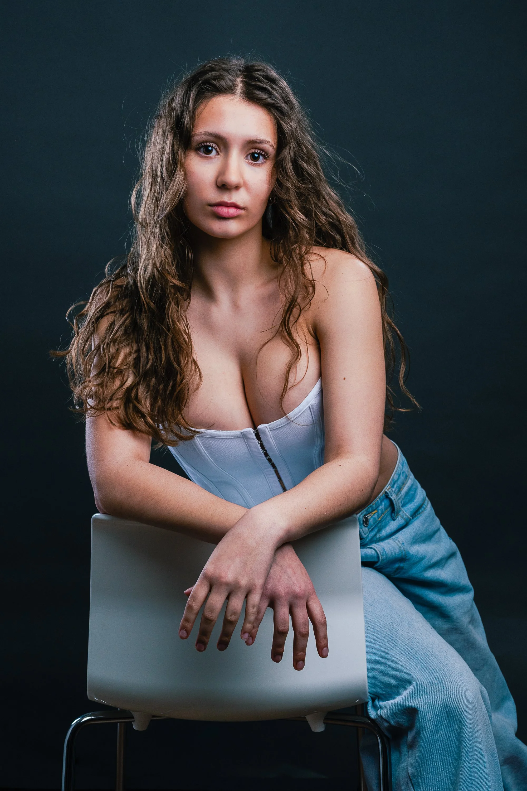A young woman with long, curly brown hair posing against a dark background. She is wearing a white corset top and light blue jeans, and is sitting on a white chair, leaning forward with her right arm resting on the chair back.