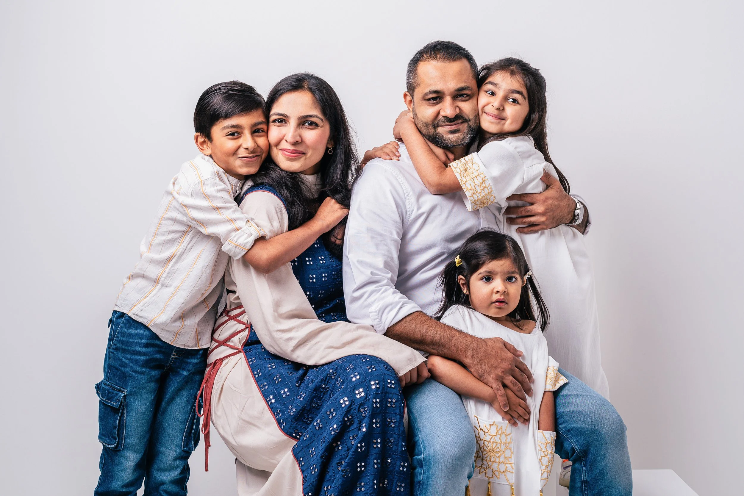 A family of six, including two adults and four children, posing together against a plain white background. The children are hugging their parents and all are smiling or looking at the camera.