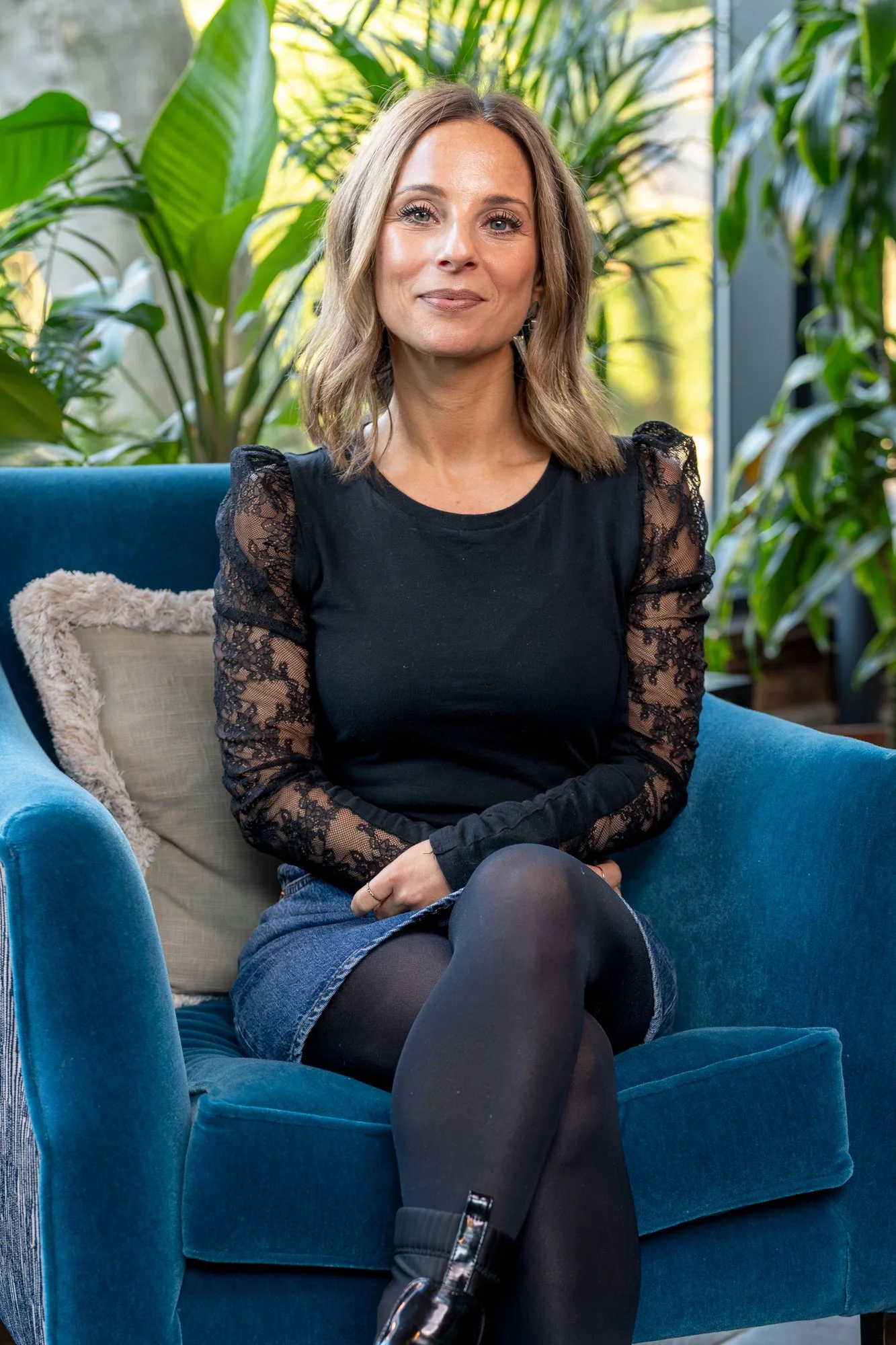 A woman with shoulder-length brown hair, wearing a black lace long-sleeve top and a denim skirt, sitting on a blue armchair with a white pillow, surrounded by lush green plants.