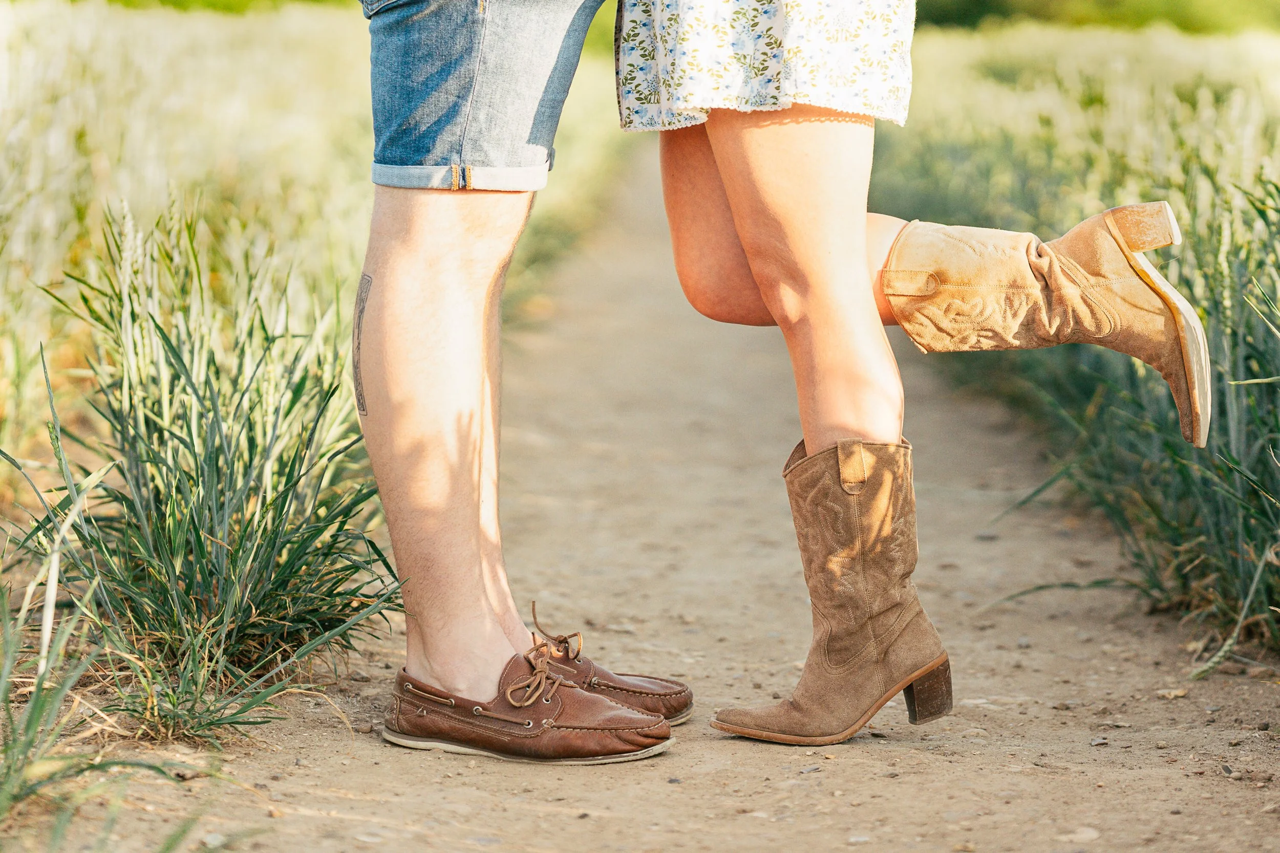 Two people standing on a dirt path in a field, with one person wearing cowboy boots and the other wearing moccasin shoes.