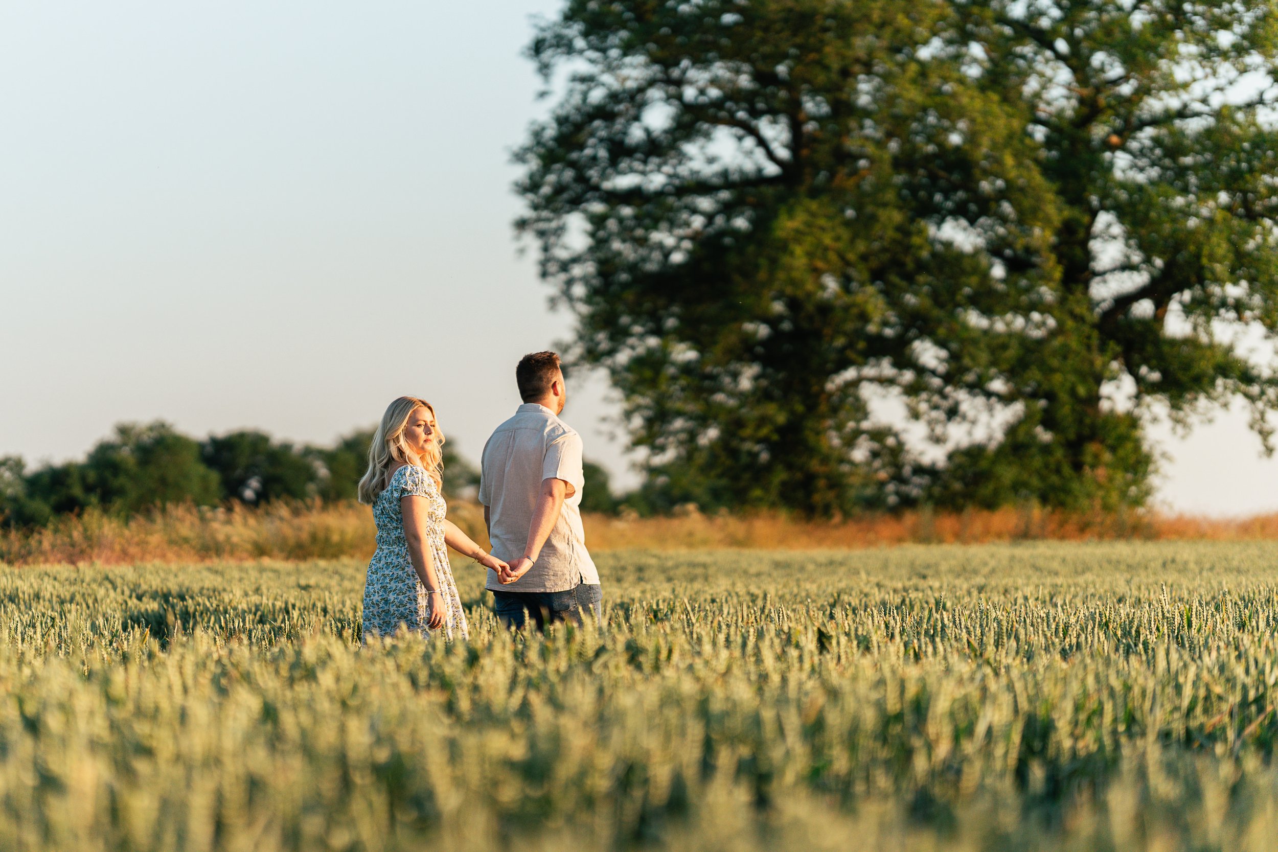 A couple holding hands walking through a wheat field with a large tree in the background, during sunset.