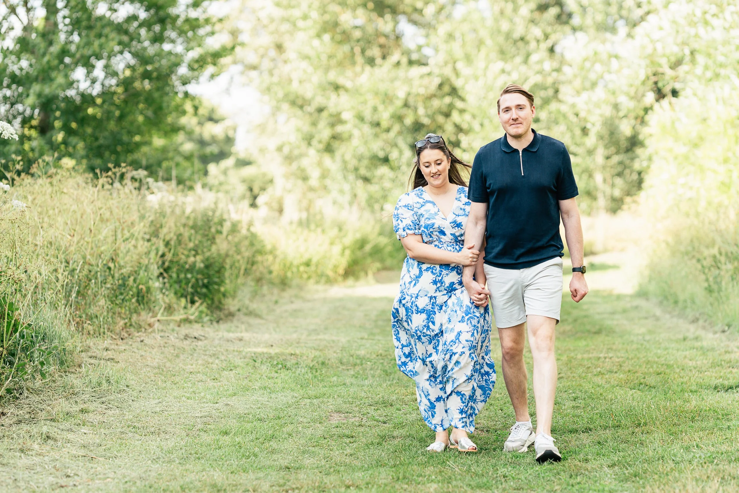 A young couple walking hand in hand on a grassy path surrounded by green trees and foliage on a sunny day.
