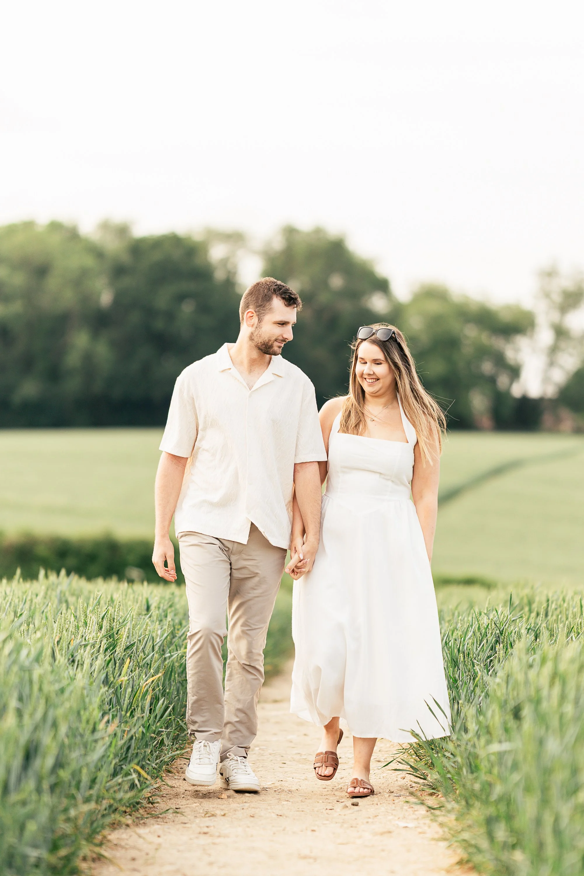 A young couple walking hand in hand on a dirt path through a green field, smiling and enjoying each other's company.