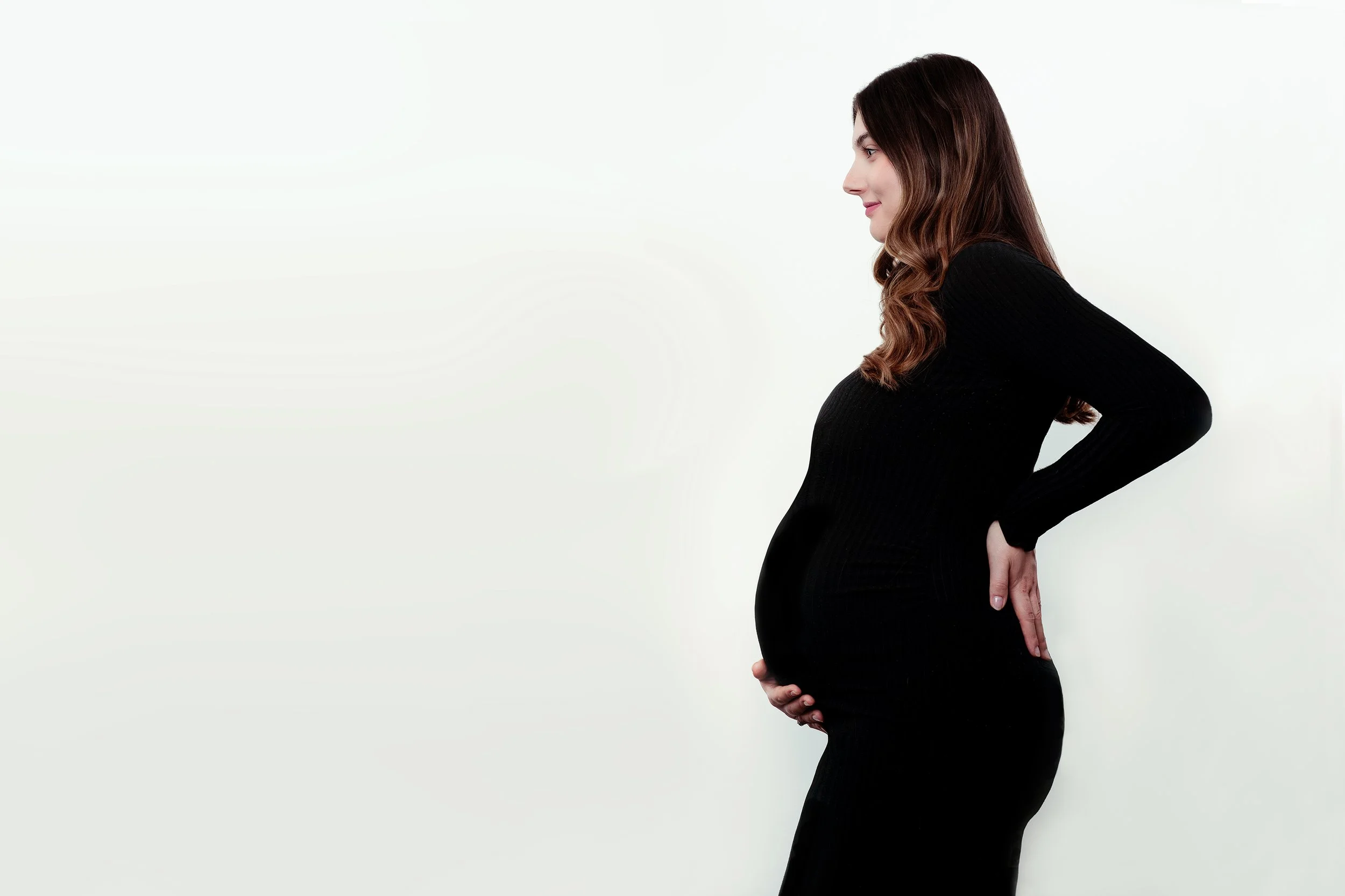 Profile of a pregnant woman in a black long-sleeve dress, standing against a white wall, with hands on her lower back and her belly.
