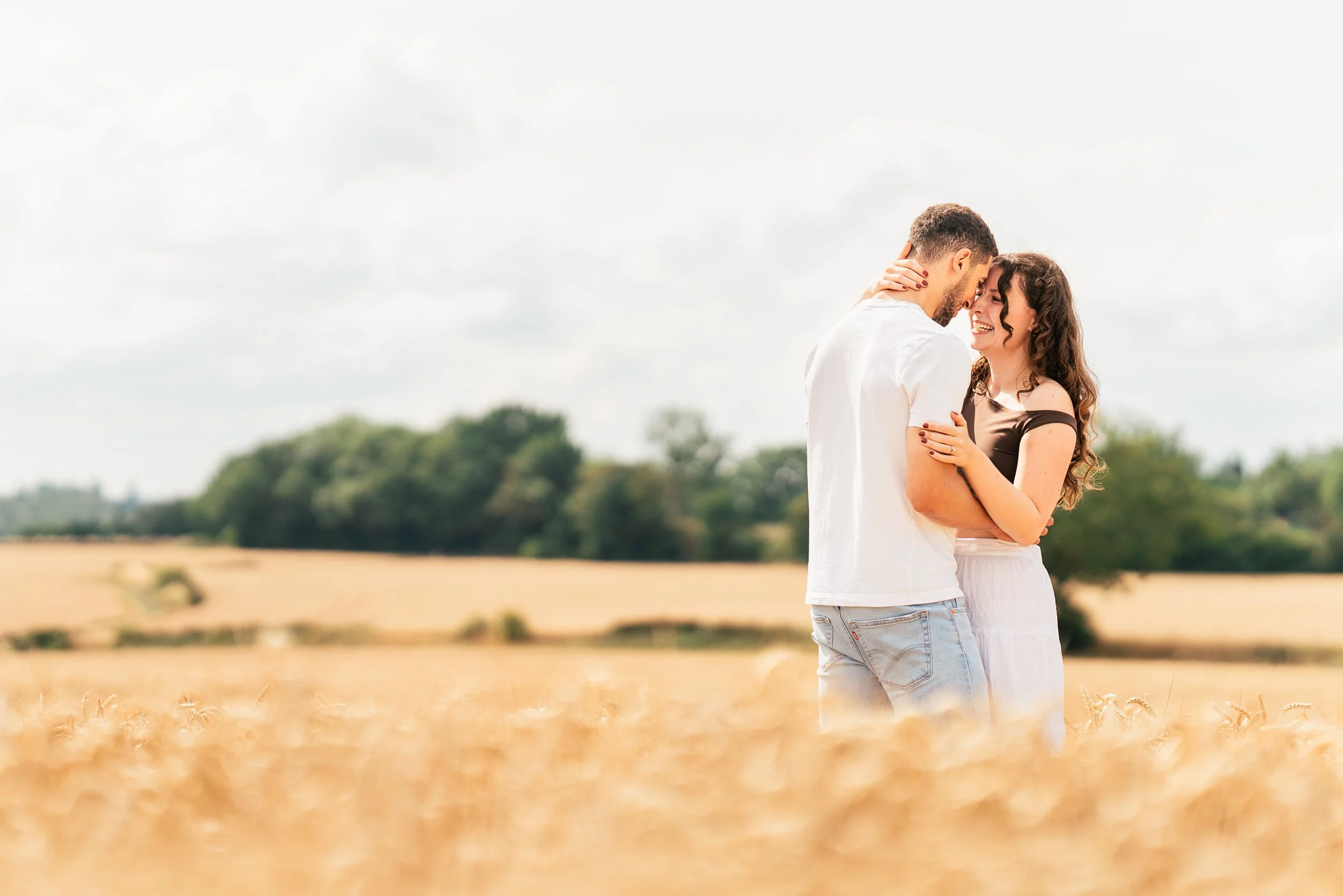 A man and a woman standing close together in a wheat field, smiling and appearing happy, with a background of trees and a cloudy sky.