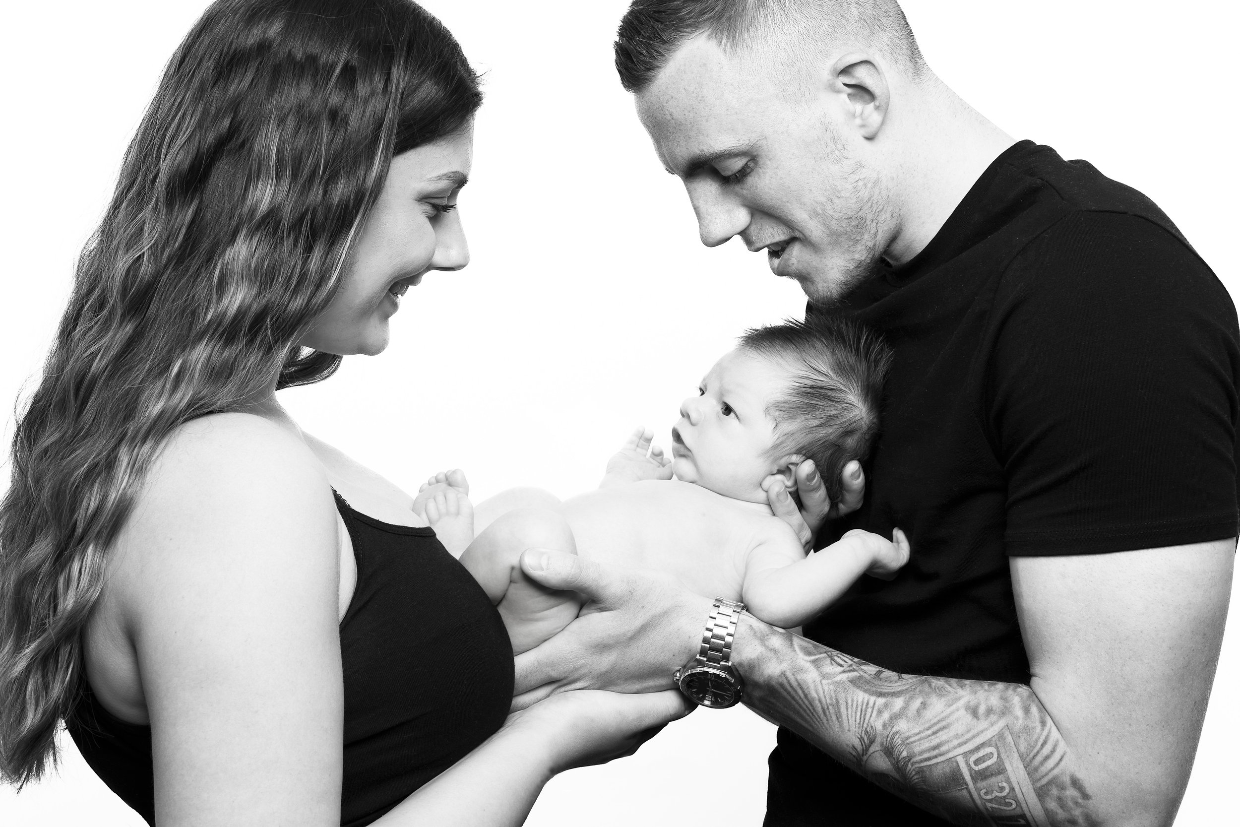 A happy woman and man holding a newborn baby together, smiling and looking at each other with love, in a black-and-white photo.