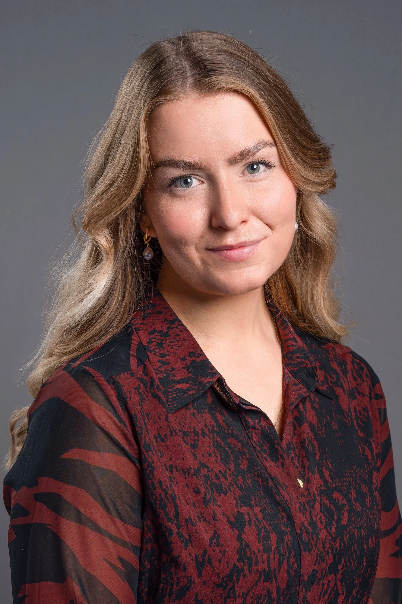 A woman with wavy blonde hair wearing a dark red and black patterned blouse, smiling against a neutral gray background.