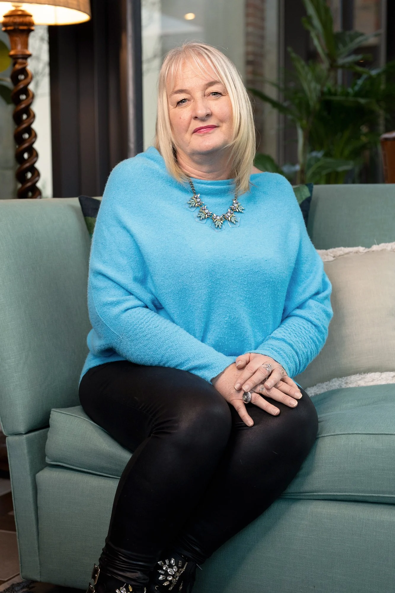 A woman with blonde hair sitting on a light green sofa, wearing a blue sweater, black pants, and jewelry, with a background of large windows and indoor plants.
