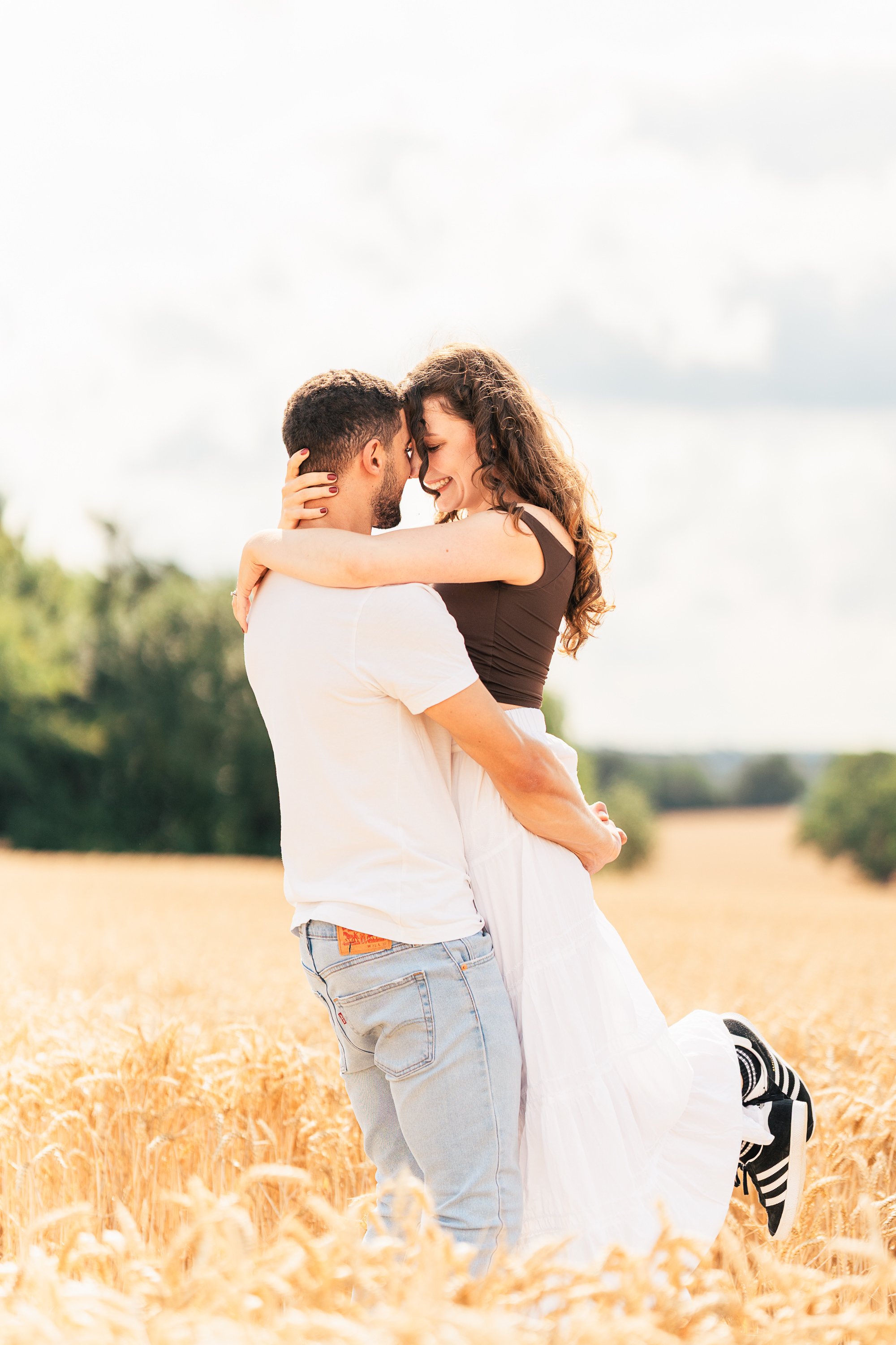 A couple embracing in a wheat field, smiling with their foreheads touching, during daytime with a partly cloudy sky.