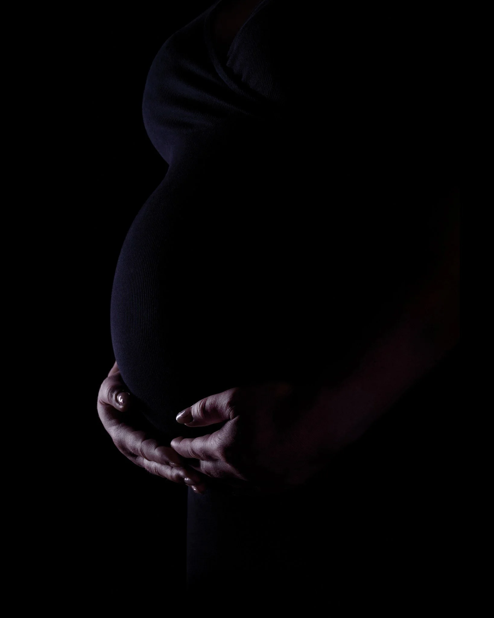 A pregnant woman wearing a black top, holding her belly with both hands in a dark setting.