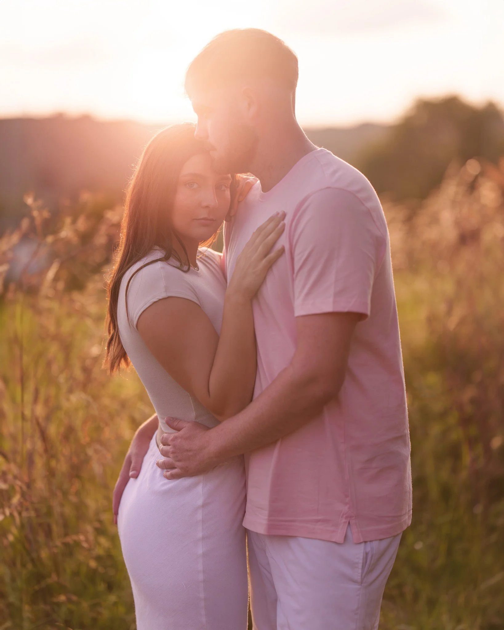 A couple standing close in a field during sunset with the man gently holding the woman's waist and the woman resting her hand on his chest, both wearing light-colored clothing.