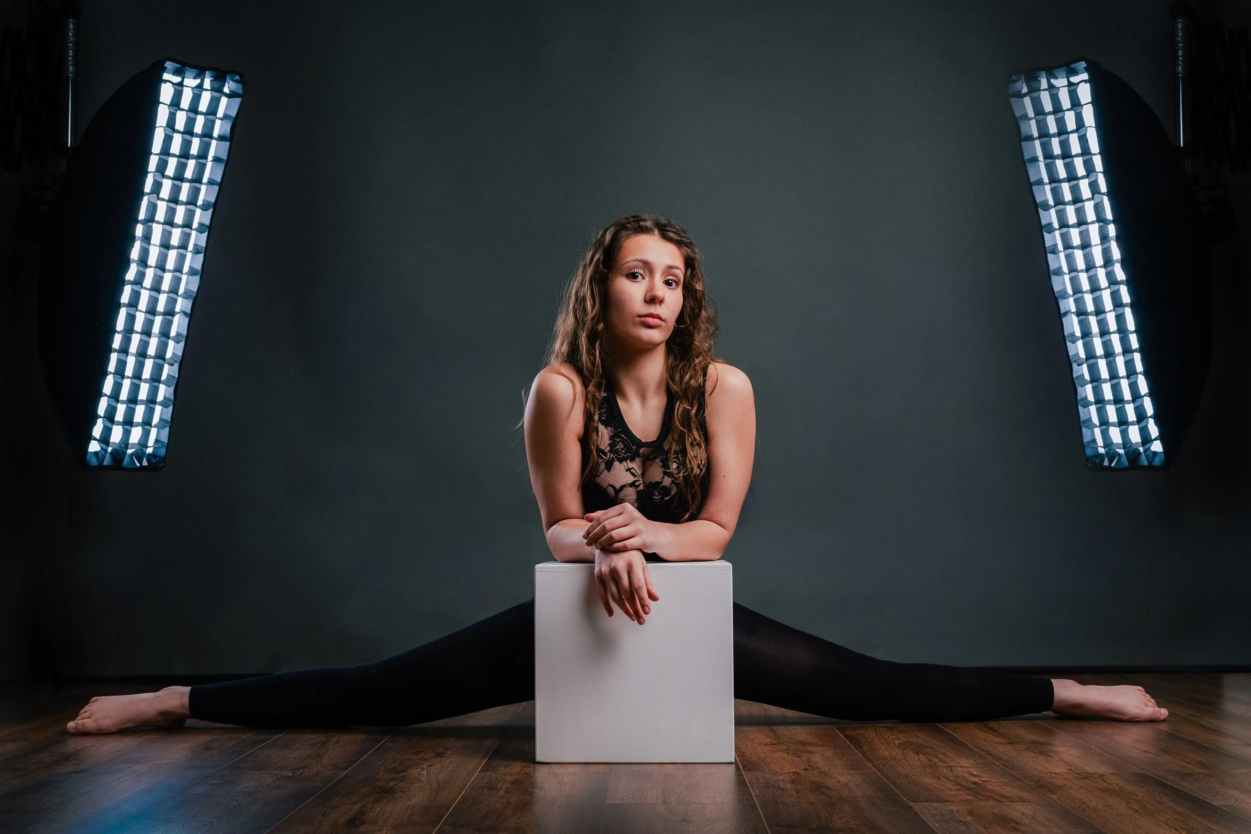 Young woman with wavy hair in a black lace top doing a split on the floor, leaning on a white box, with studio lights on either side.