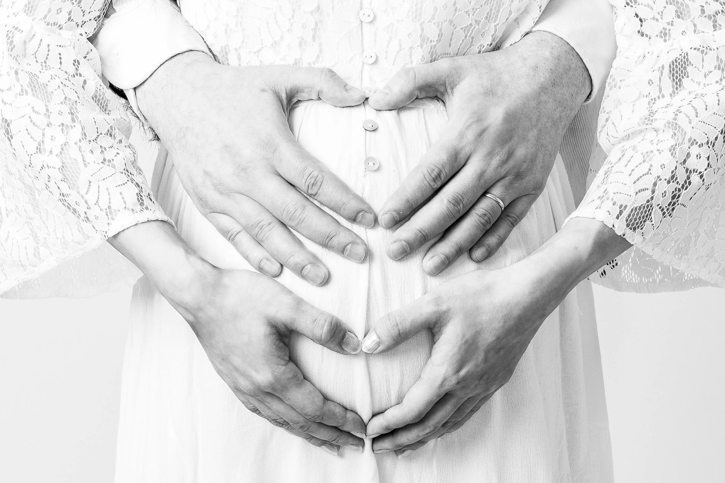 Black and white photo of a pregnant woman with her hands on her belly, forming a heart shape with two pairs of hands around her stomach.