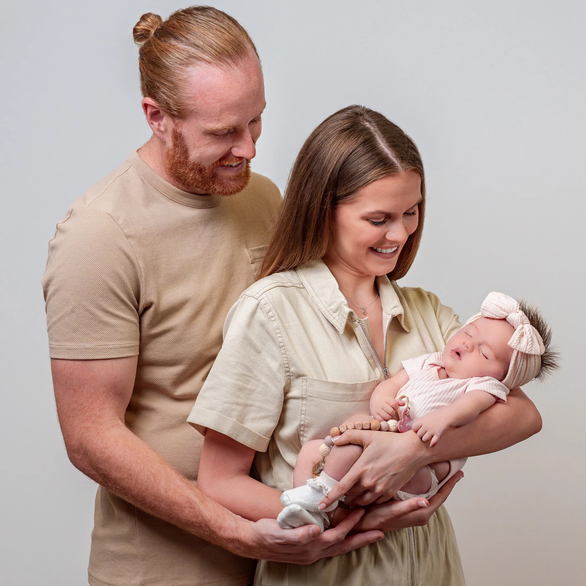 A woman holding a sleeping baby with a man standing beside her, both smiling, against a plain light-colored background.