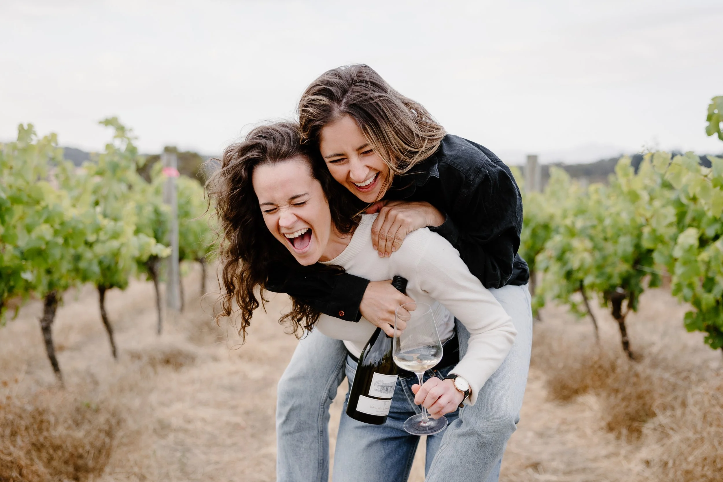 The third generation of Shepherd's Hut vineyard in Western Australia's Great Southern wine region. Two women authentically laughing and play in the Porongurup vineyard, looking over the Stirling Range. One holds a wine bottle and glass of Riesling.