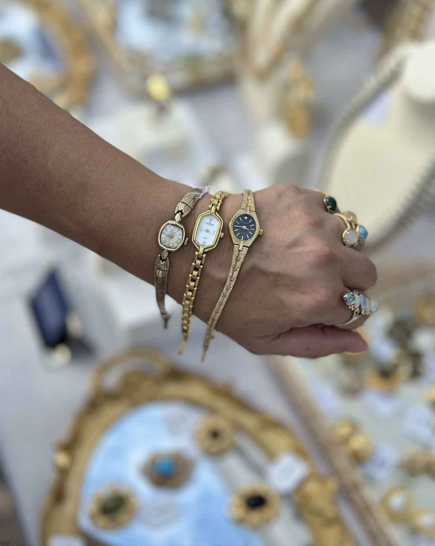 Close-up of a person's wrist wearing multiple gold and silver watches and rings, with a jewelry display in the blurred background.