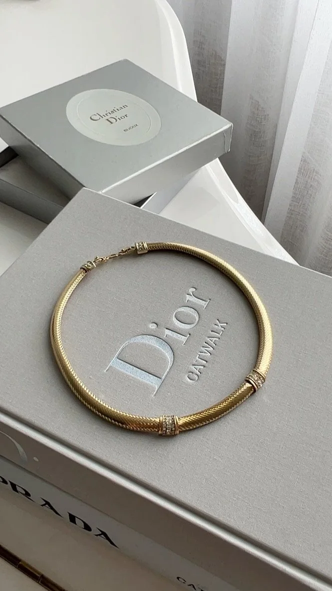 A gold necklace with diamond accents resting on a gray Christian Dior box, with additional Christian Dior packaging in the background.