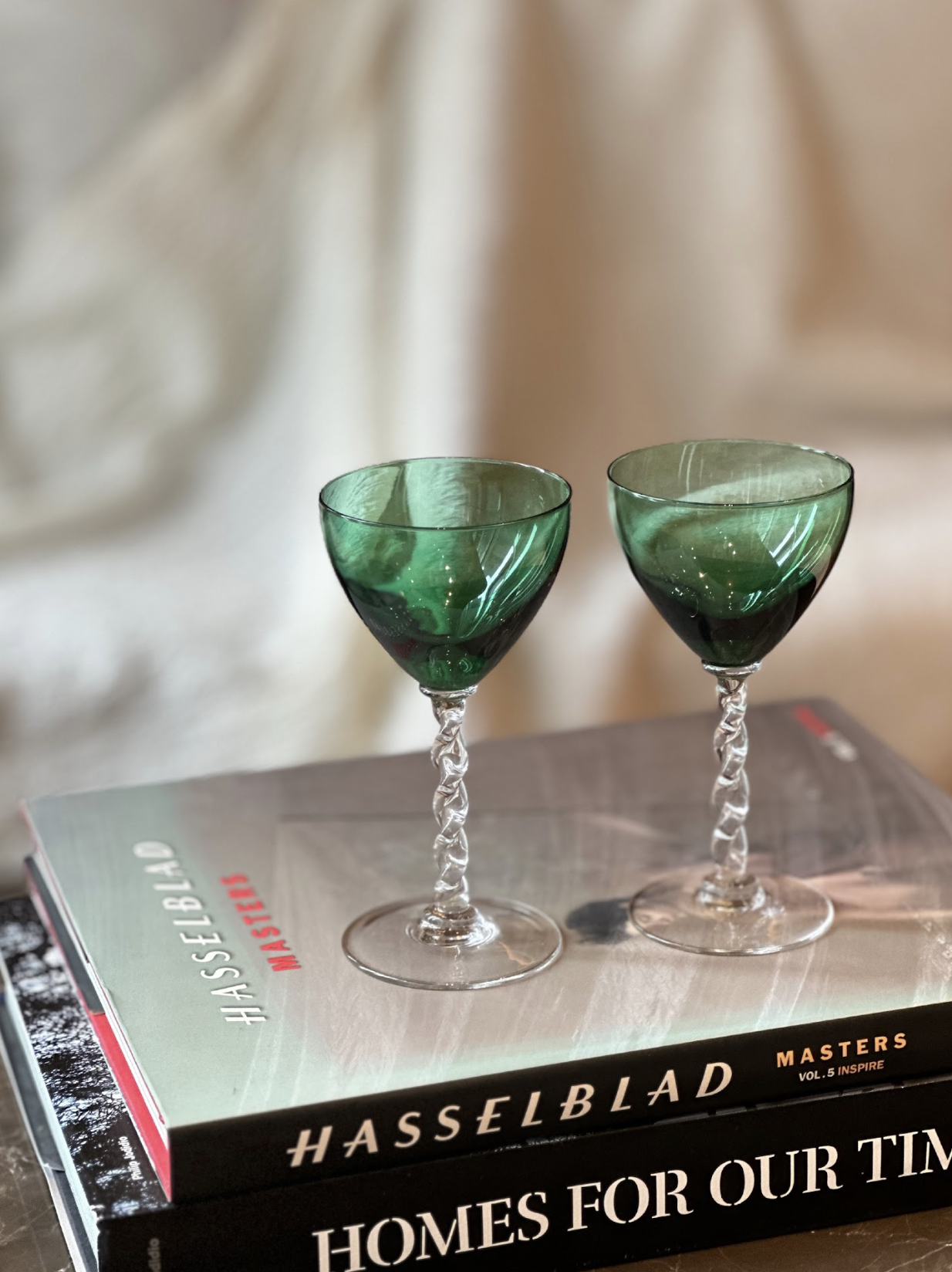 Two green glass wine goblets with twisted stems on top of a stack of books, with a blurred beige background.
