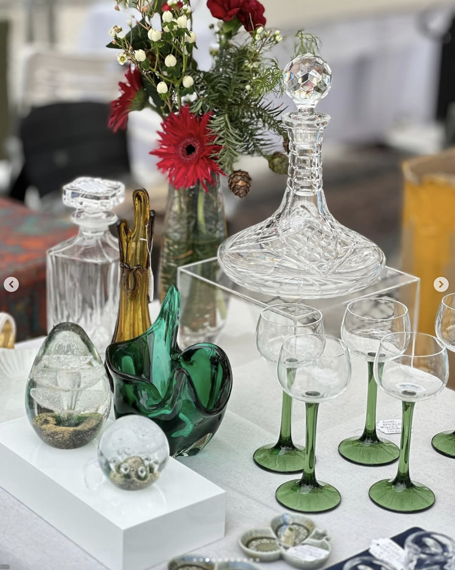 A display of various decorative glassware including vases, decanters, and glasses, with a background of a floral arrangement in a jar and other items on a table.