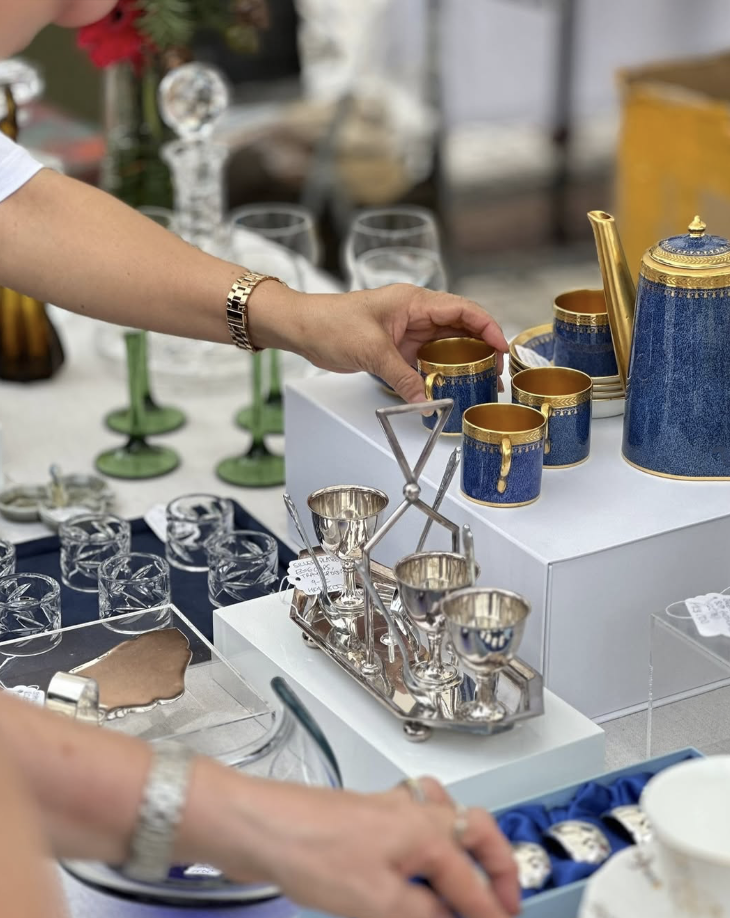 Person holding a blue and gold tea set on display at a market or store, with various glassware and decorative items visible on the table.