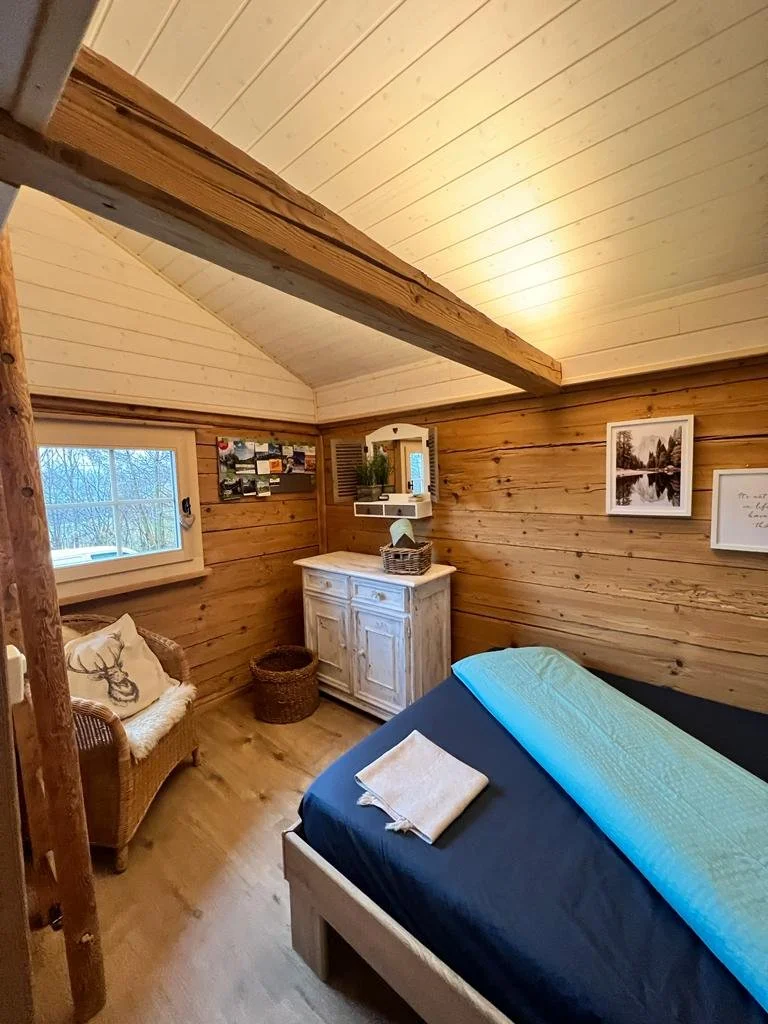 Kaminzimmer mit Holzwänden und steiler Decke, Bett mit blauer Decke, Korb, Nistkasten, Bilder an der Wand und Fenster mit Aussicht auf Bäume im Winter.
