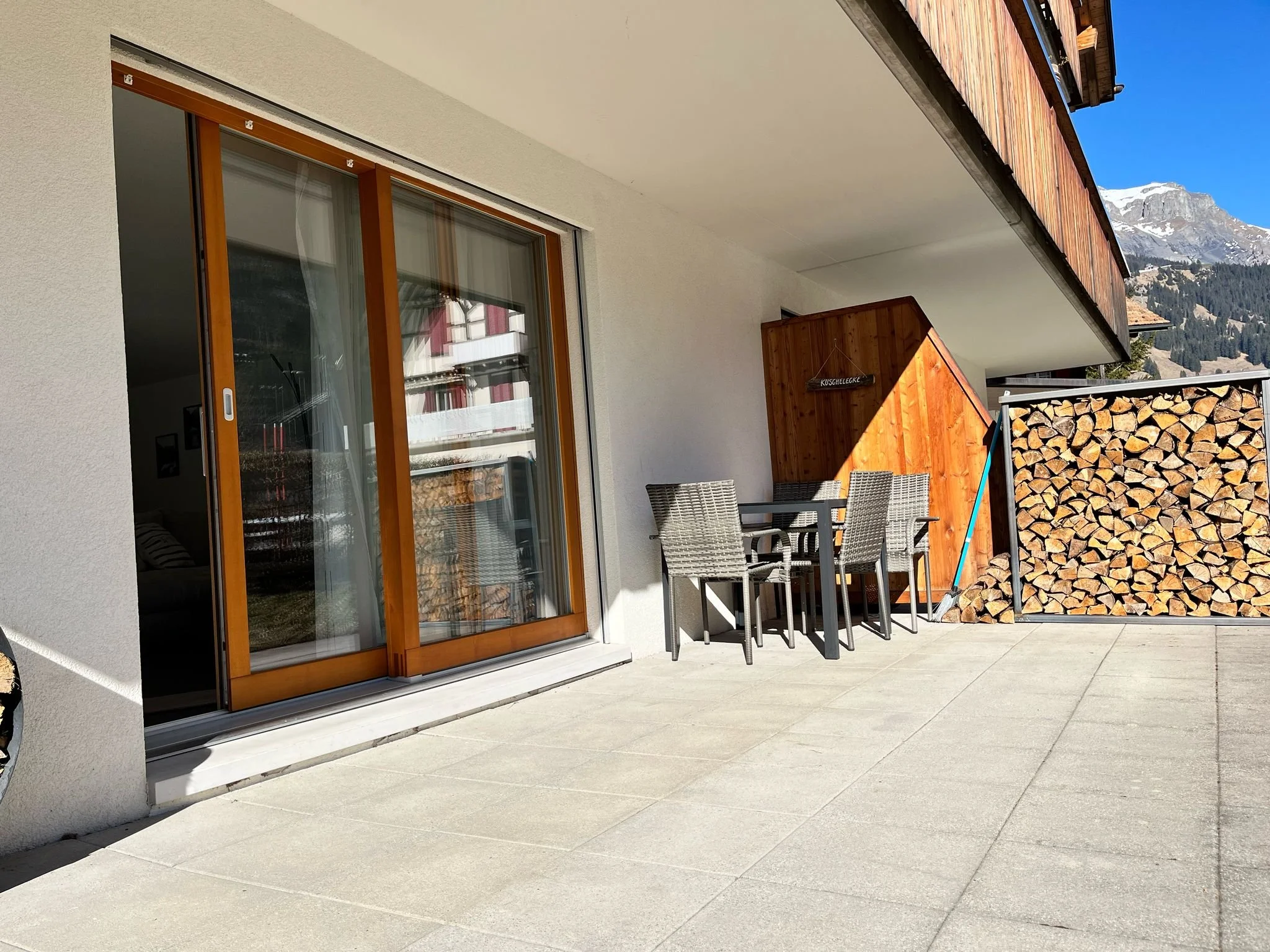 Balkon mit Sitzgruppe, Holzscheiten, Holzhaus, Berge im Hintergrund, sonnig, heller Himmel.