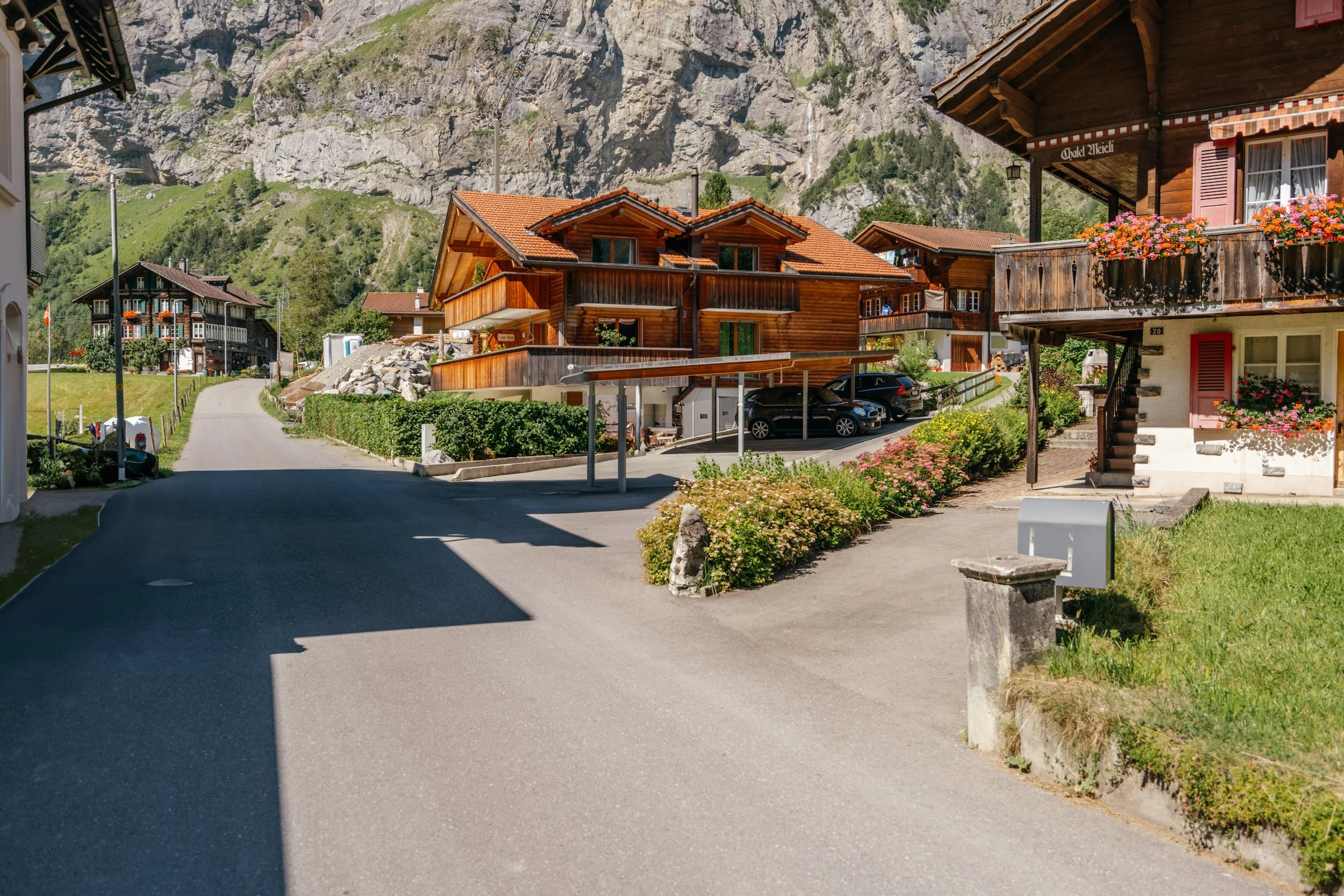 Wohnhäuser in einer alpinen Bergregion, mit Holzfassaden, blühenden Blumen und einer asphaltierte Straße im Vordergrund bei Sonnenlicht.