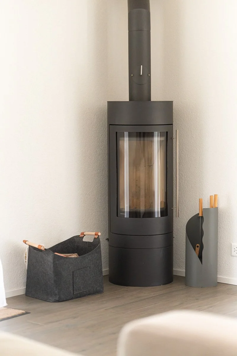 Ein moderner, schwarze Kaminofen vor einer weißen Wand mit einem Kaminbesteck und einem Aufbewahrungsbehälter daneben.
